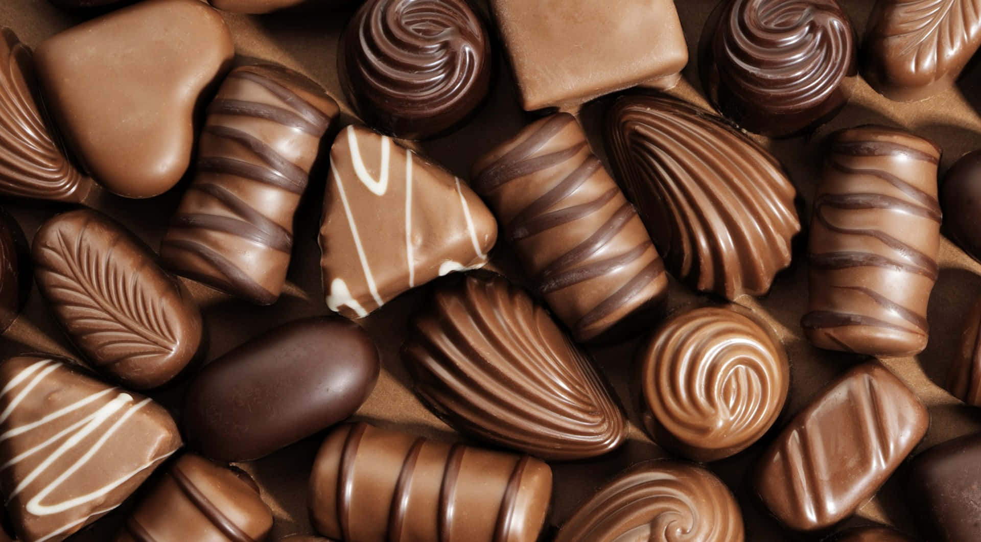 Brown Chocolate 桌布 壁紙 — 各種光澤巧克力的特寫，帶有漩渦、條紋和葉子圖案，呈現豐富的棕色色調。