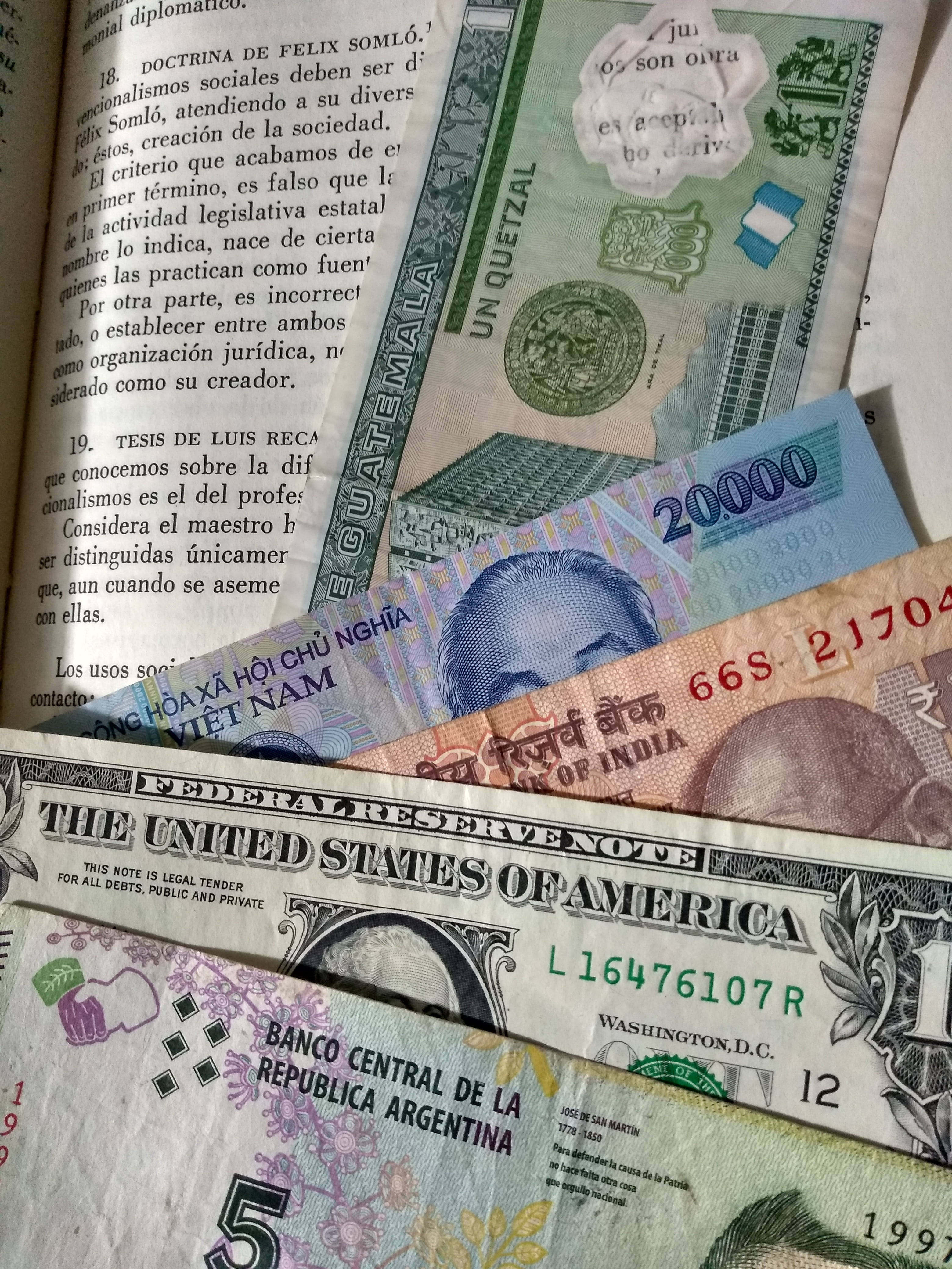 money iPhone 桌布 壁紙 — 各種全球紙幣的特寫，包括美元、印度盧比和瓜地馬拉格查爾，疊放在一本帶有西班牙語文字的打開的書上。
