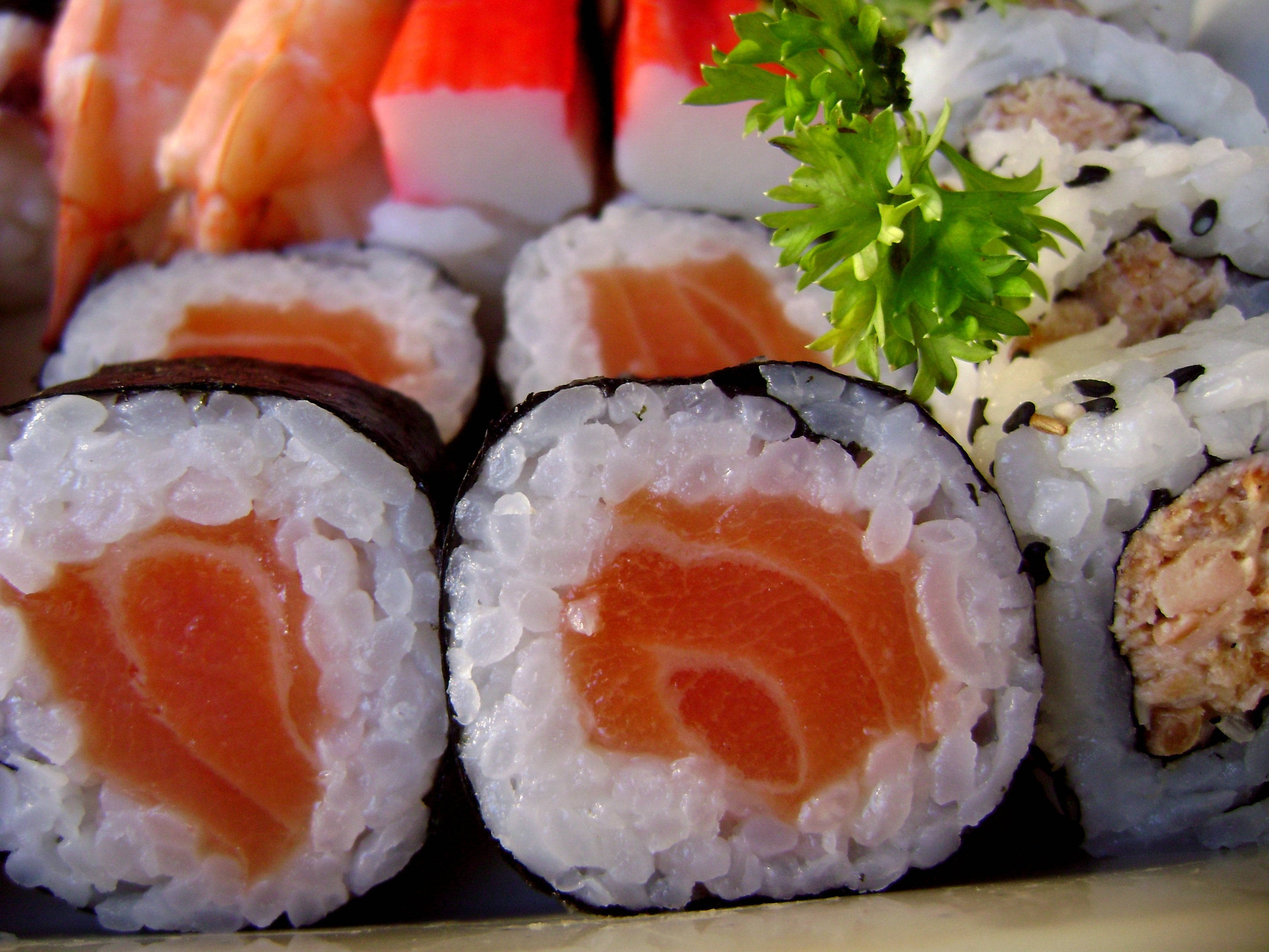 Sushi 桌布 壁紙 — 各種壽司卷和握壽司的特寫，展示新鮮的鮭魚、鮪魚、蝦和蟹，並以新鮮歐芹裝飾。