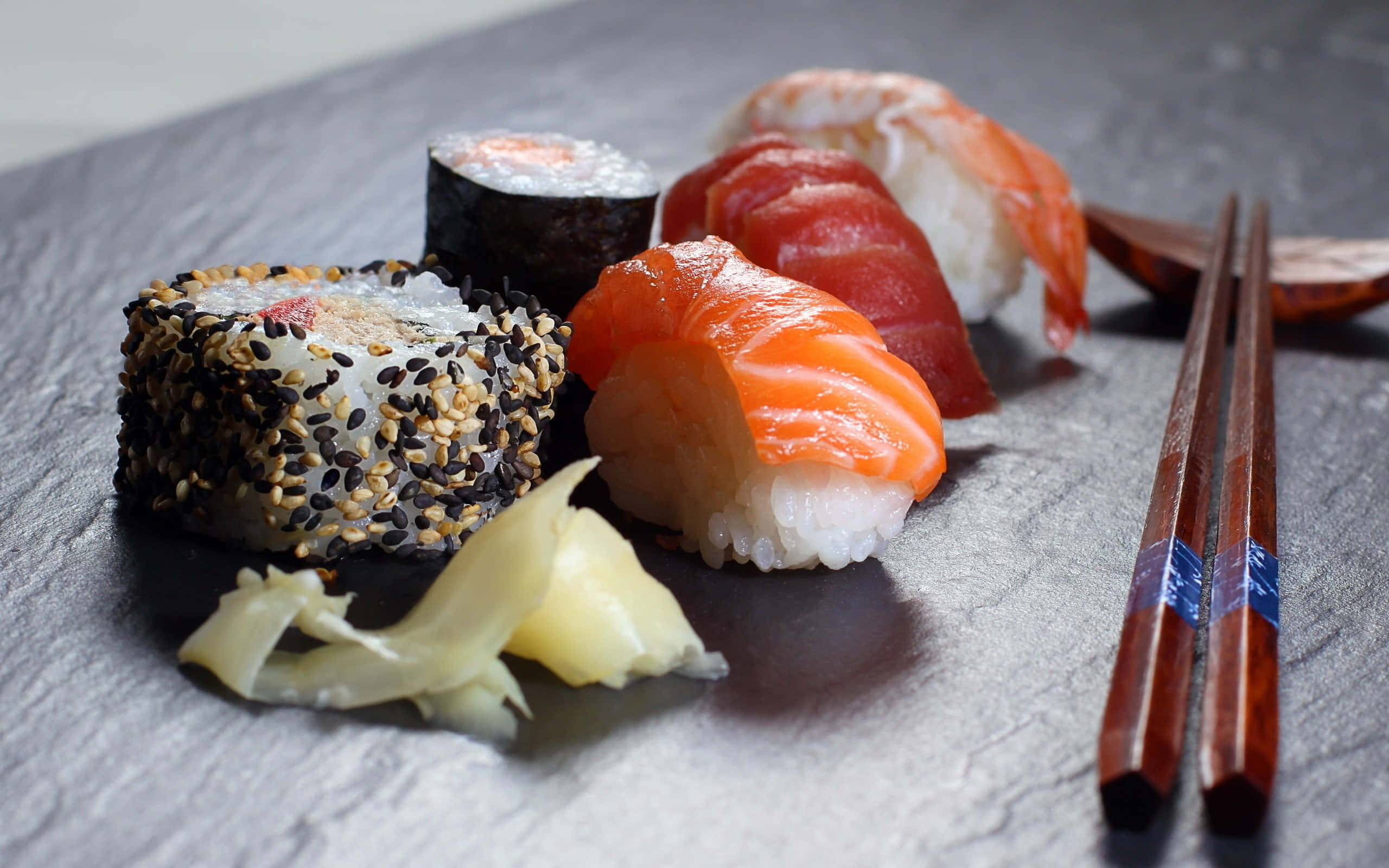 Sushi 桌布 壁紙 — 各種壽司拼盤的特寫，包括鮭魚握壽司和芝麻卷，擺放在深色石板上的筷子與芥末。
