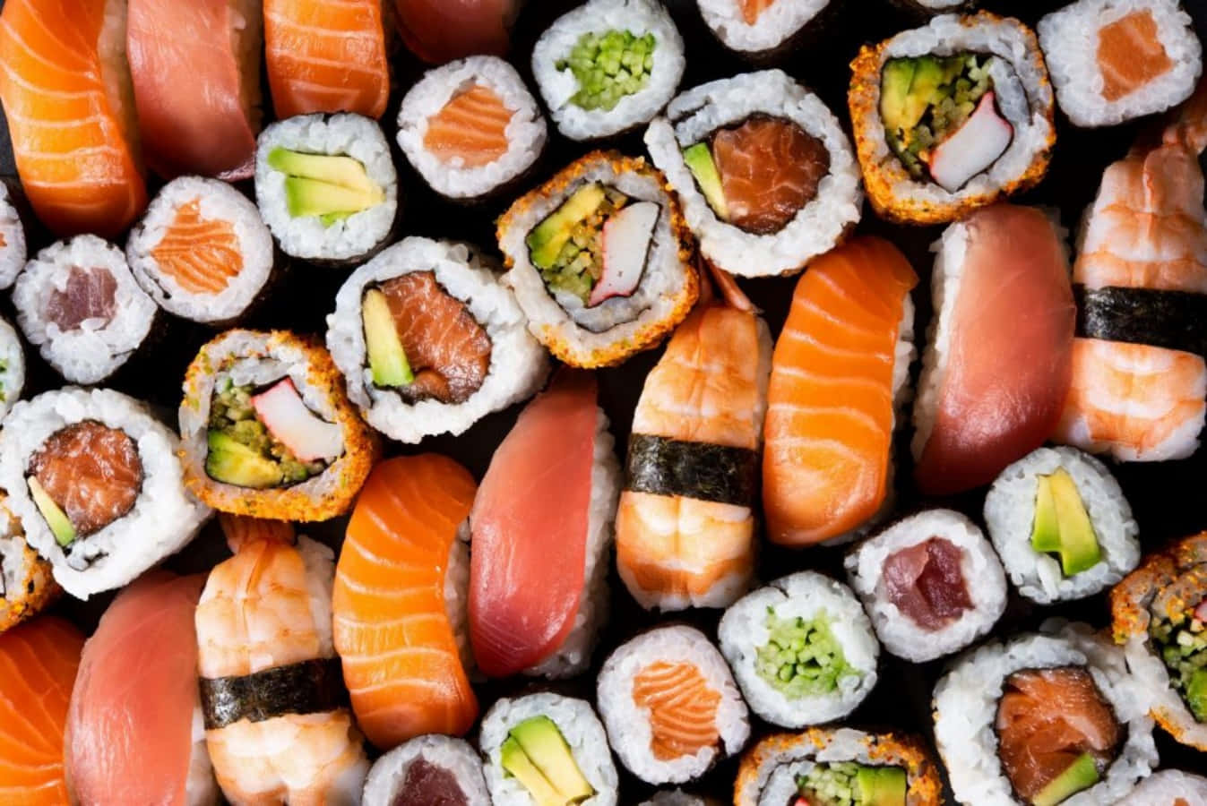 壽司 桌布 壁紙 — 各種壽司的鮮豔特寫，包括鮭魚、鮪魚、酪梨和小黃瓜卷，密集排列在深色表面上。