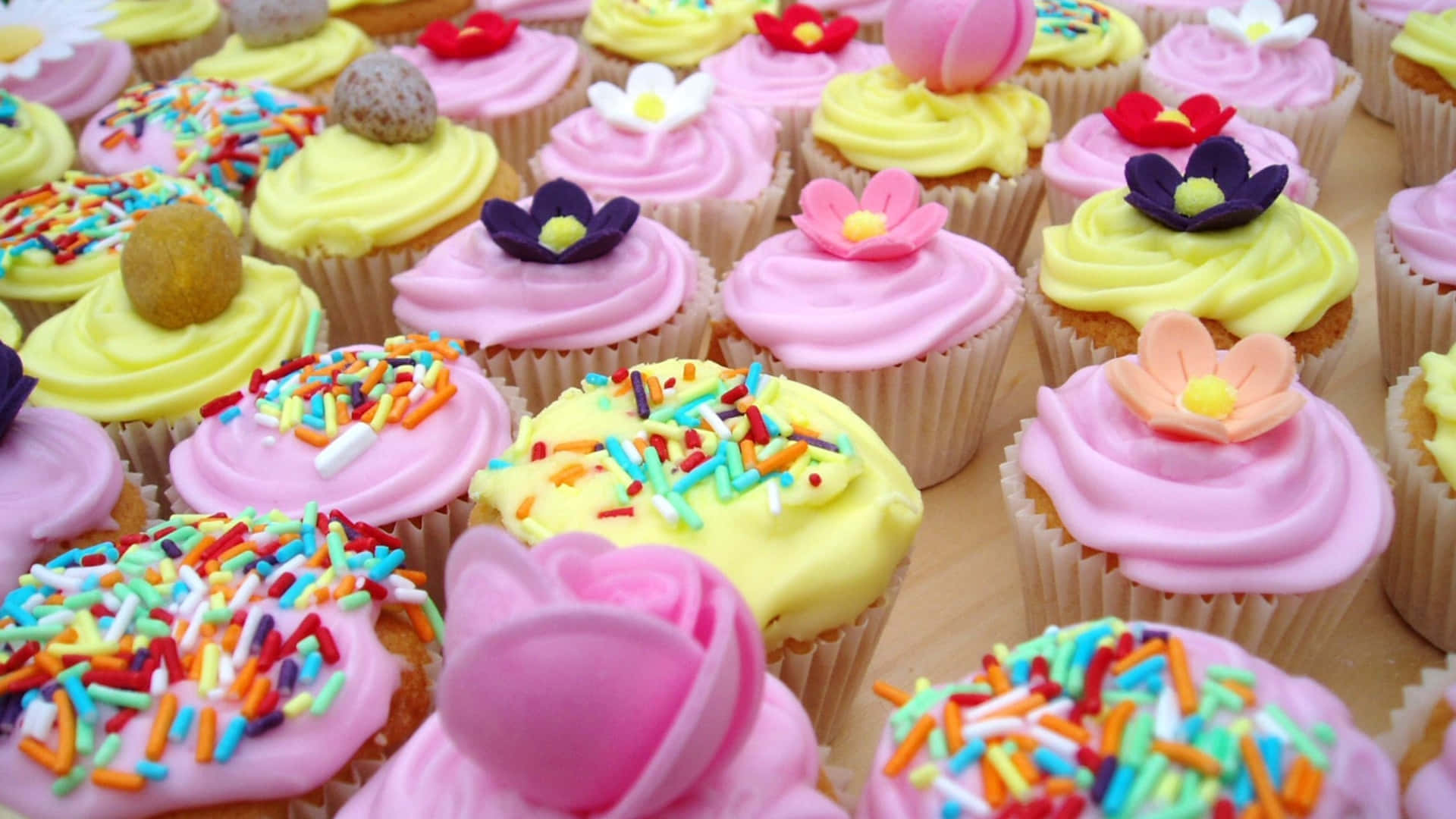 Cupcake 桌布 壁紙 — 各種杯子蛋糕的特寫，上面有粉紅色和黃色的糖霜、彩色糖粒和可食用花朵。