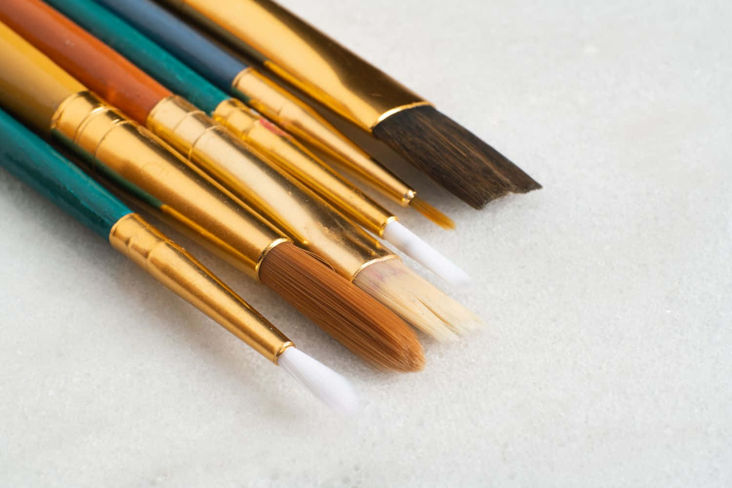 Paint Brush 桌布 壁紙 — 各種畫筆的特寫，帶有金色金屬箍和多種刷毛，放置在帶有紋理的白色表面上。