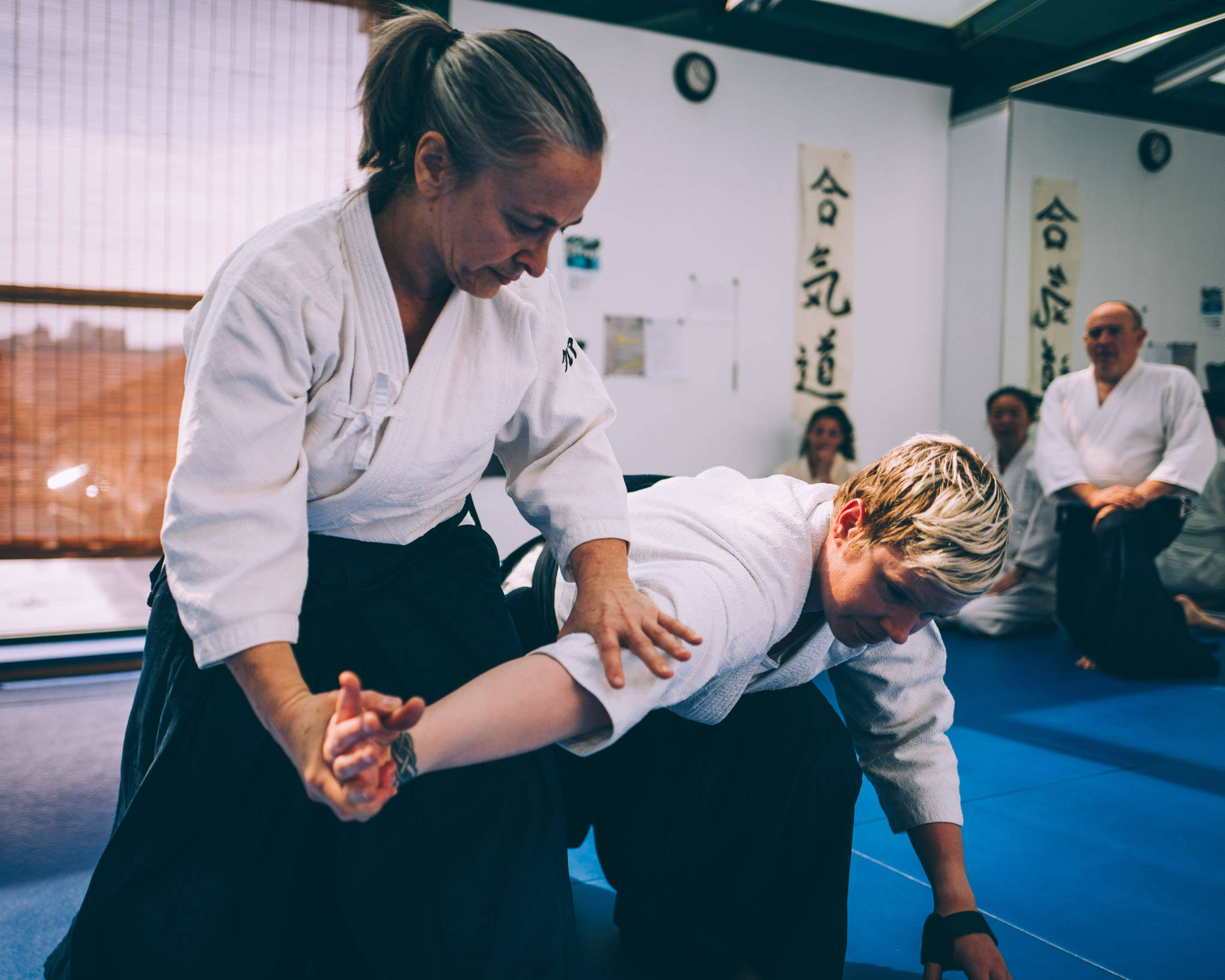 martial arts 桌布 壁紙 — 合氣道教練在道場指導學生進行精準的投摔，道場內有藍色軟墊和牆上的日本書法。