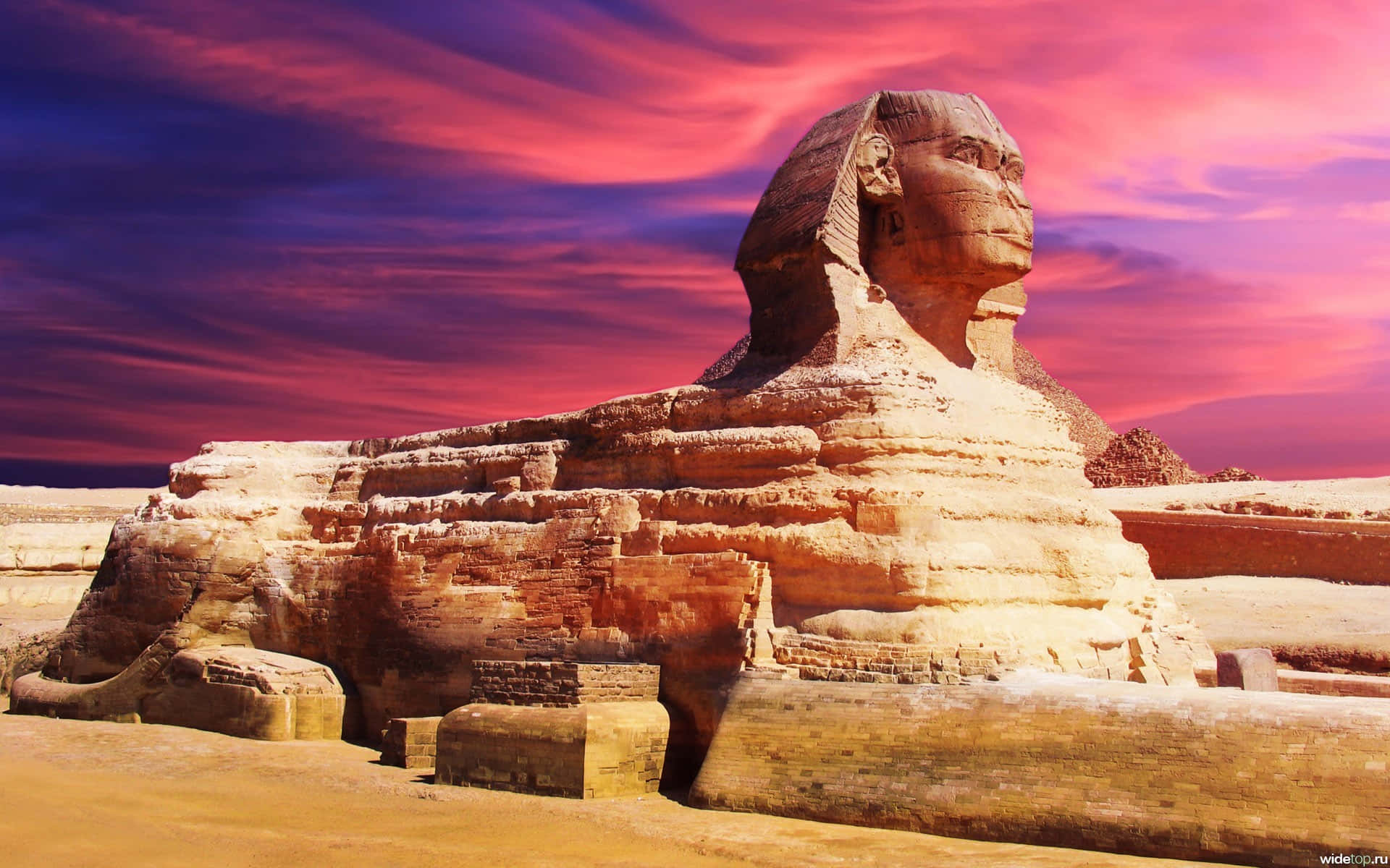 egyptian 桌布 壁紙 — 吉薩大獅身人面像在充滿活力的粉紅色和紫色日落天空下雄偉地矗立，遠處有金字塔。