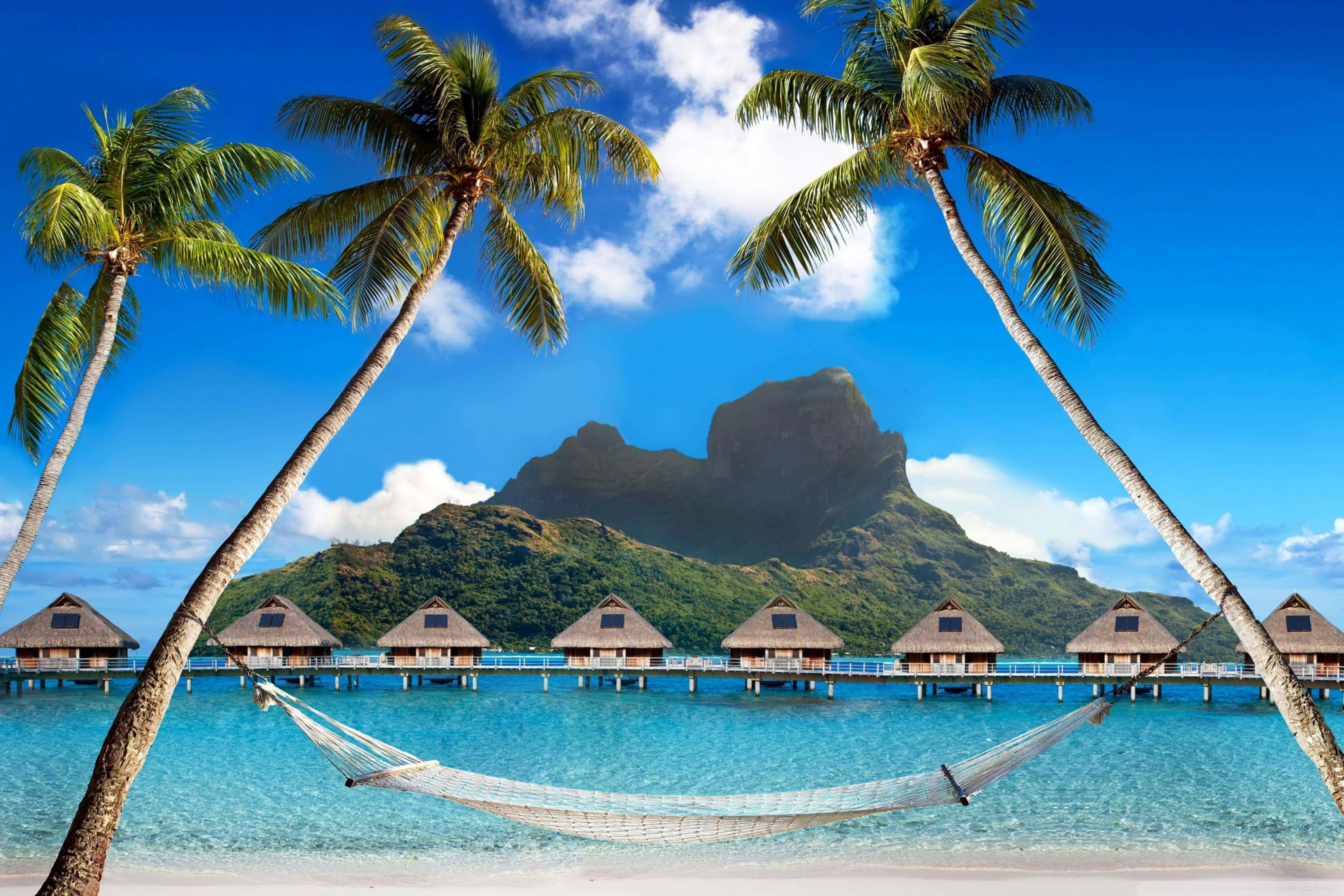 Vacation Mode 桌布 壁紙 — 吊床掛在熱帶海灘的棕櫚樹之間，背景有水上平房和鬱鬱蔥蔥的山脈，在明亮的藍天下。