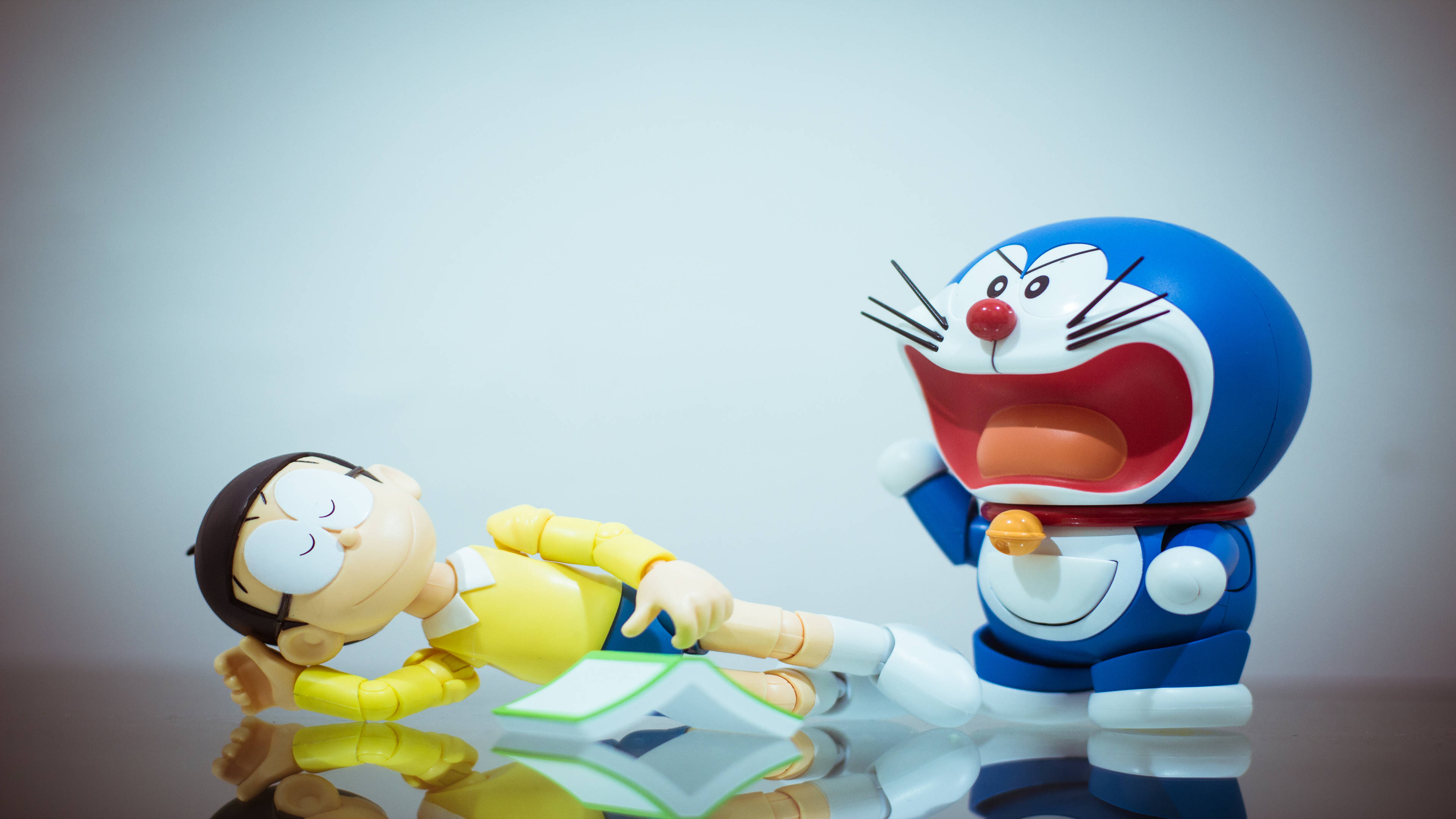 Doraemon 桌布 壁紙 — 哆啦A夢和大雄的模型在反光表面上戲劇性地擺姿勢，一個角色在笑，另一個躺著。