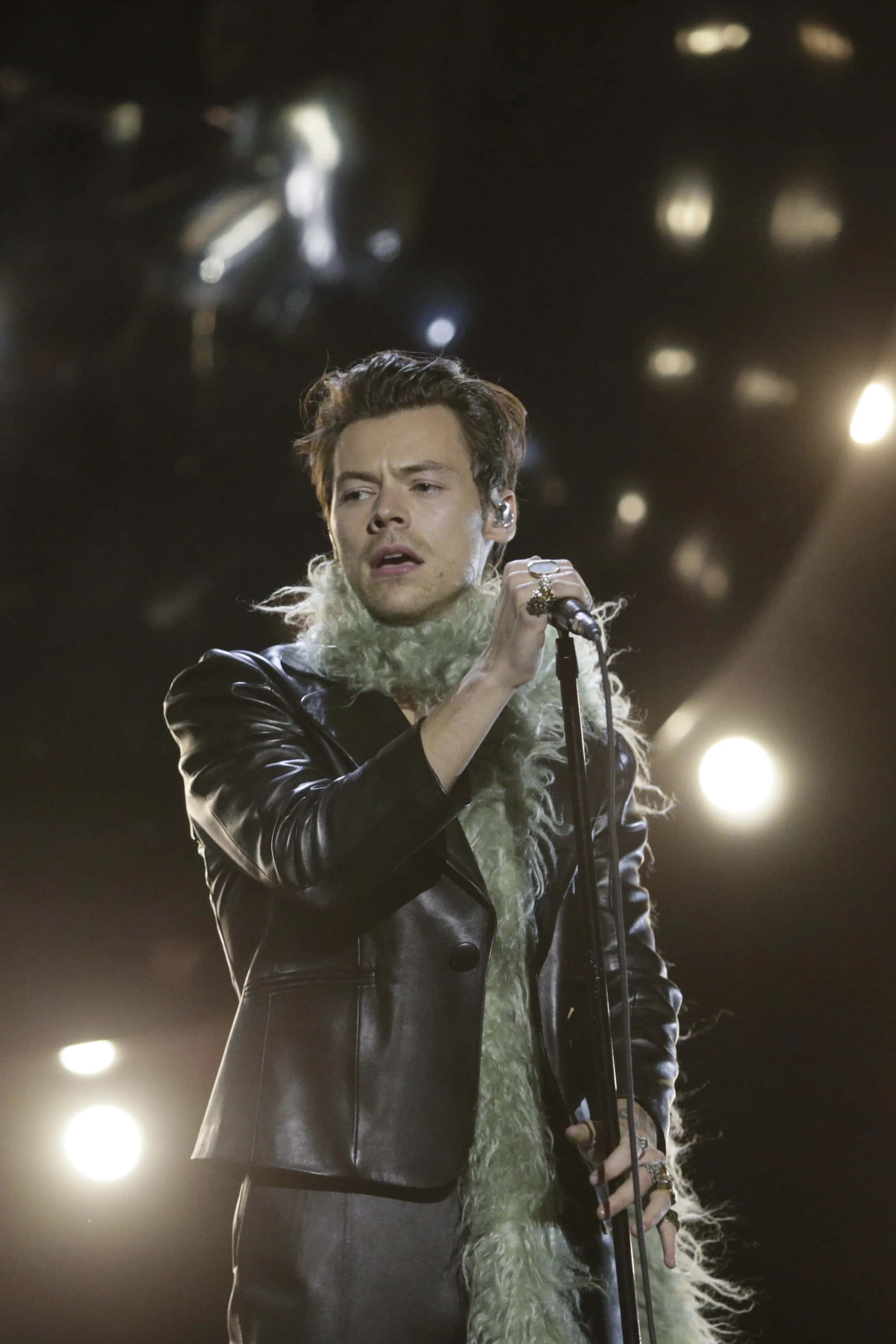 Harry Styles 2021 桌布 壁紙 — 哈利·史泰爾斯在明亮的舞台燈光下，穿著黑色皮夾克搭配綠色毛領，在舞台上激情演唱。