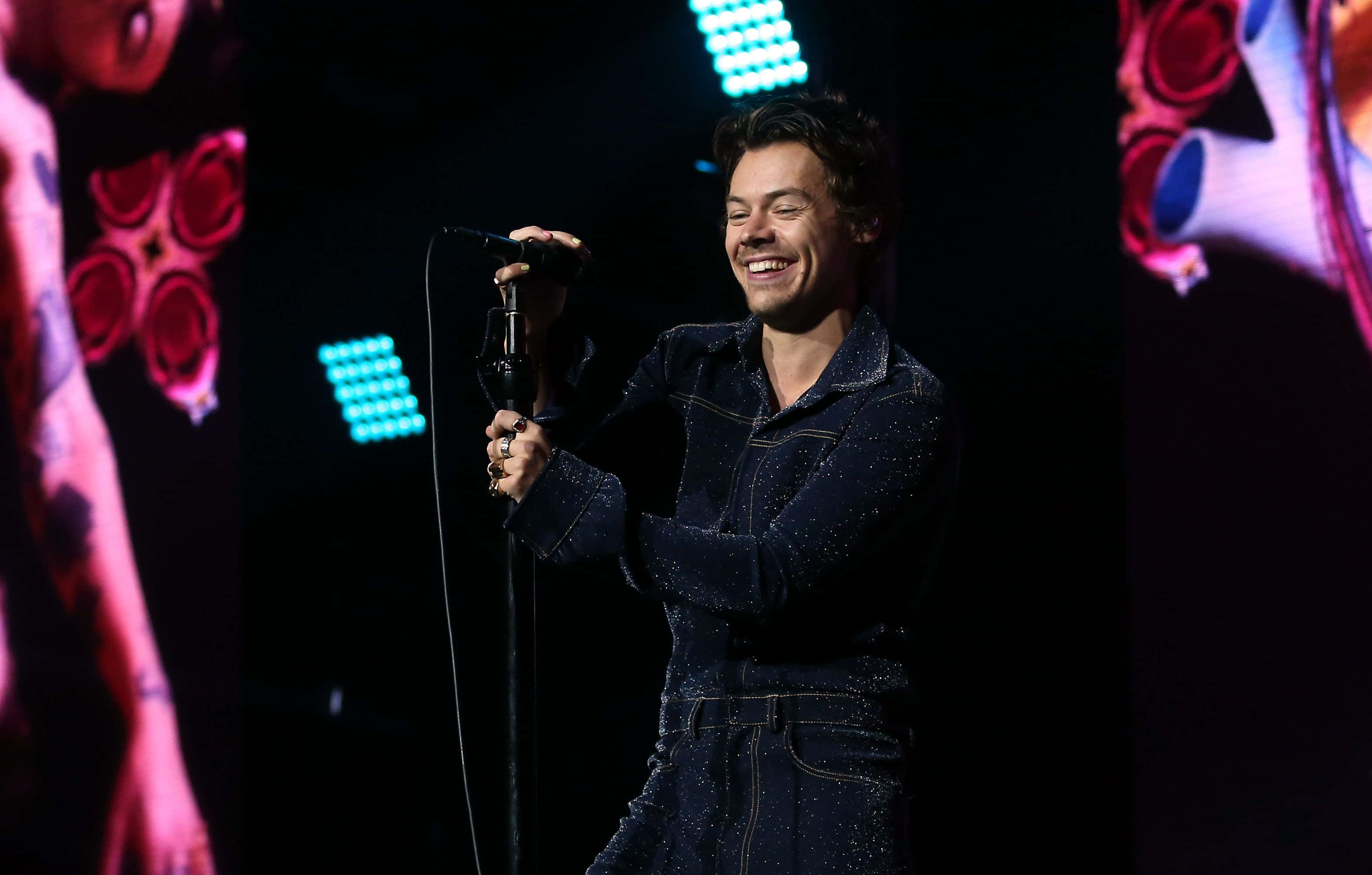 Harry Styles Laptop 桌布 壁紙 — 哈利·史泰爾斯在藍色和粉紅色燈光照耀下的活力舞台上，開心地握著麥克風，笑容燦爛。