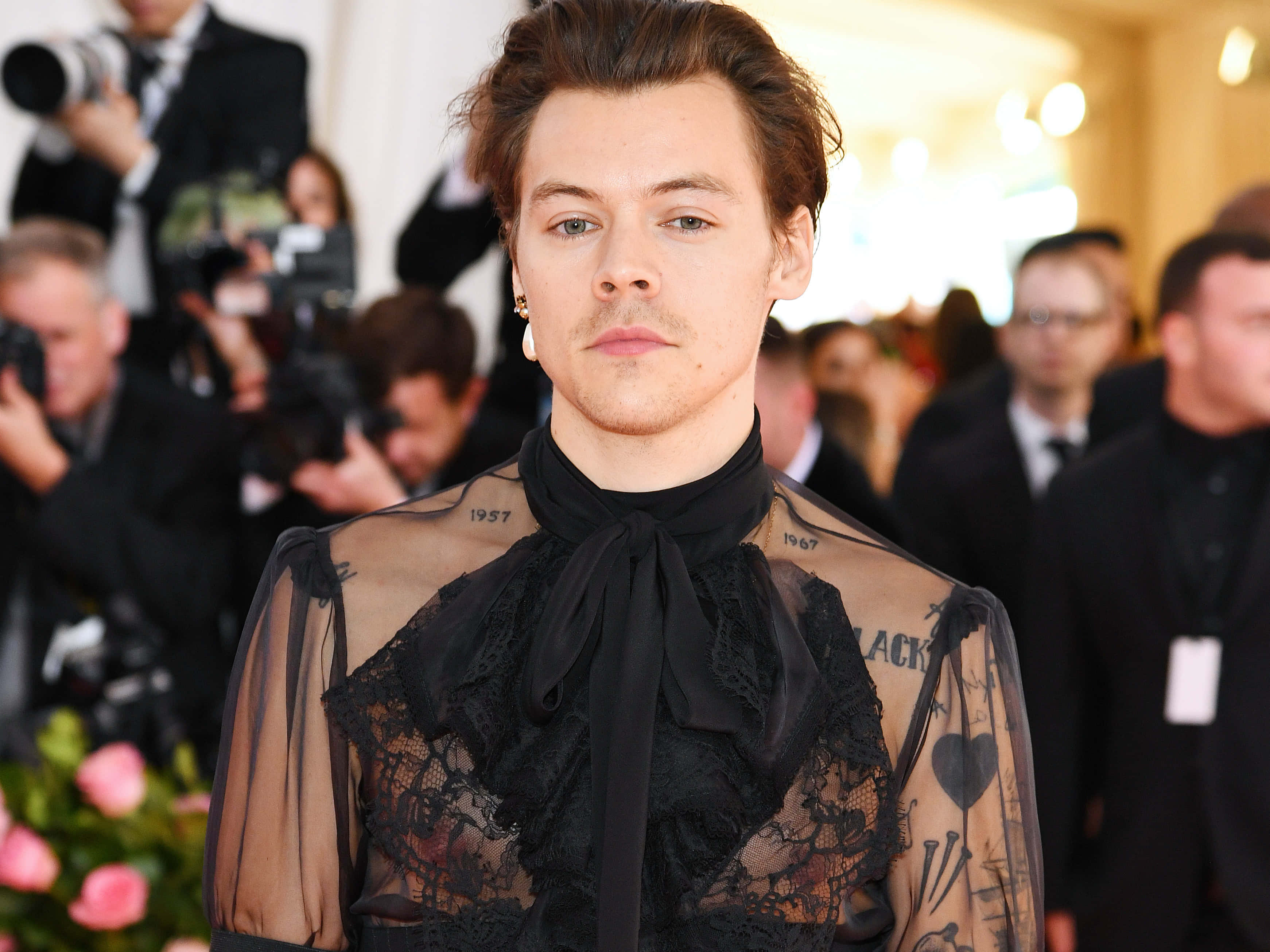 Harry Styles 2021 桌布 壁紙 — 哈利·史泰爾斯身穿透視黑色蕾絲服裝，露出可見的刺青，站在紅地毯上，被攝影師包圍。