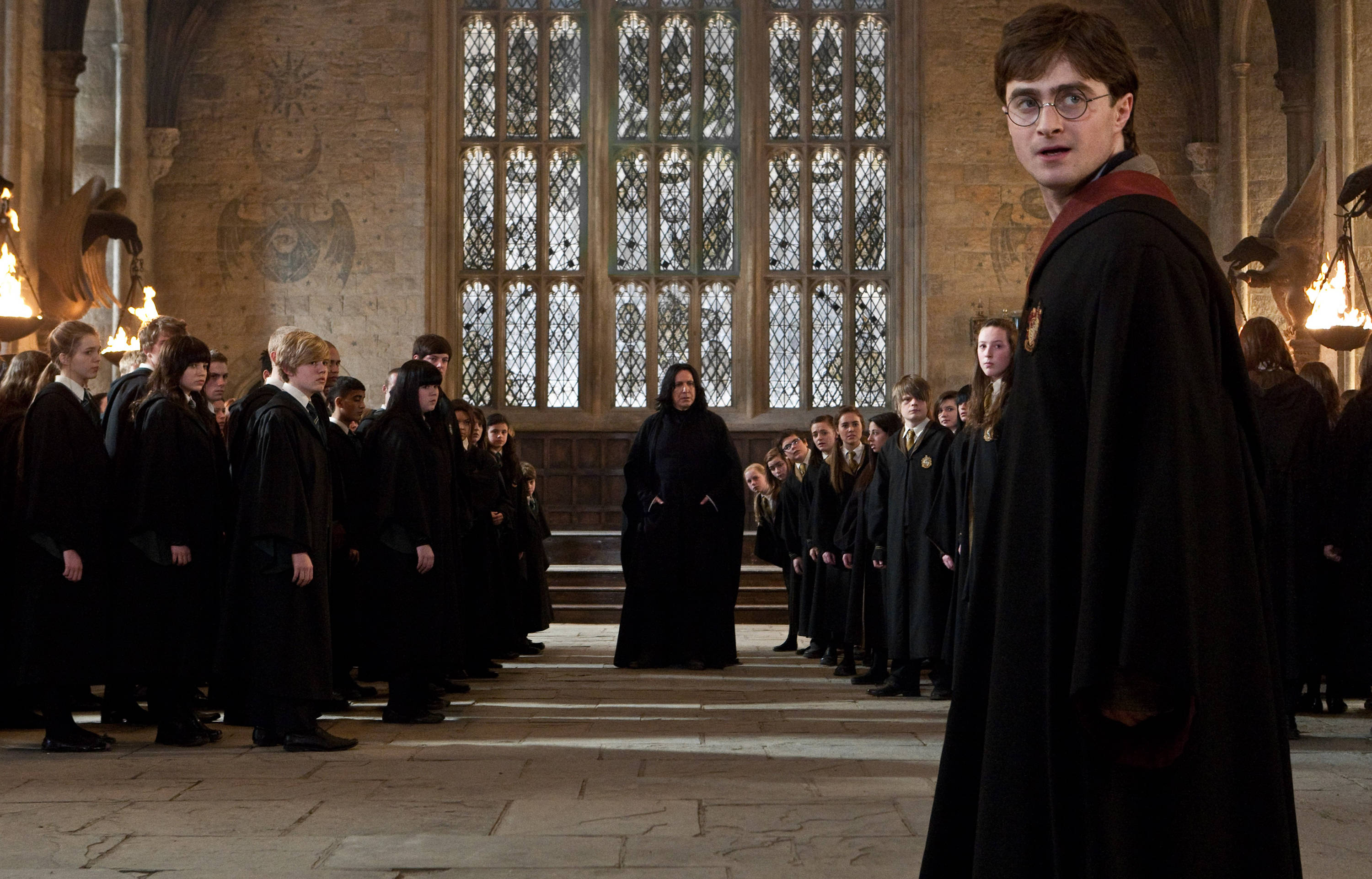Severus Snape 桌布 壁紙 — 哈利波特站在一個宏偉的石廳中，面對一位嚴厲的教授，而身穿黑色長袍的學生們排成行列。