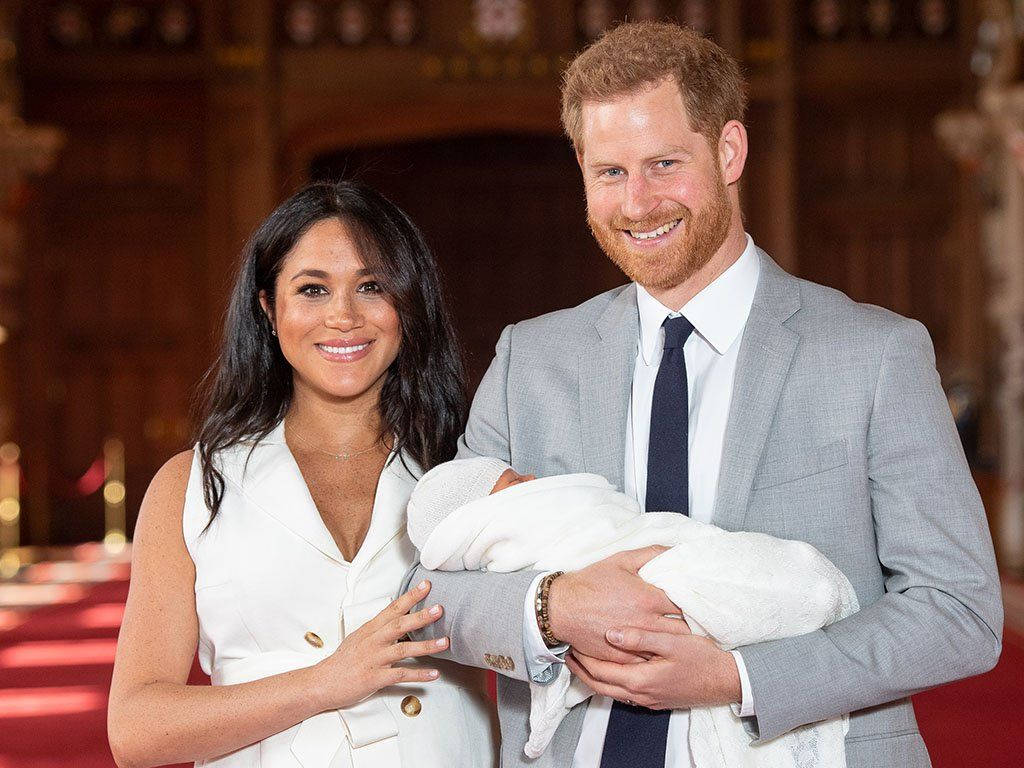 Meghan Markle 桌布 壁紙 — 哈利王子和梅根·馬克爾溫暖地微笑，抱著用白色包裹的新生嬰兒，背景是豐富的木質室內裝飾。