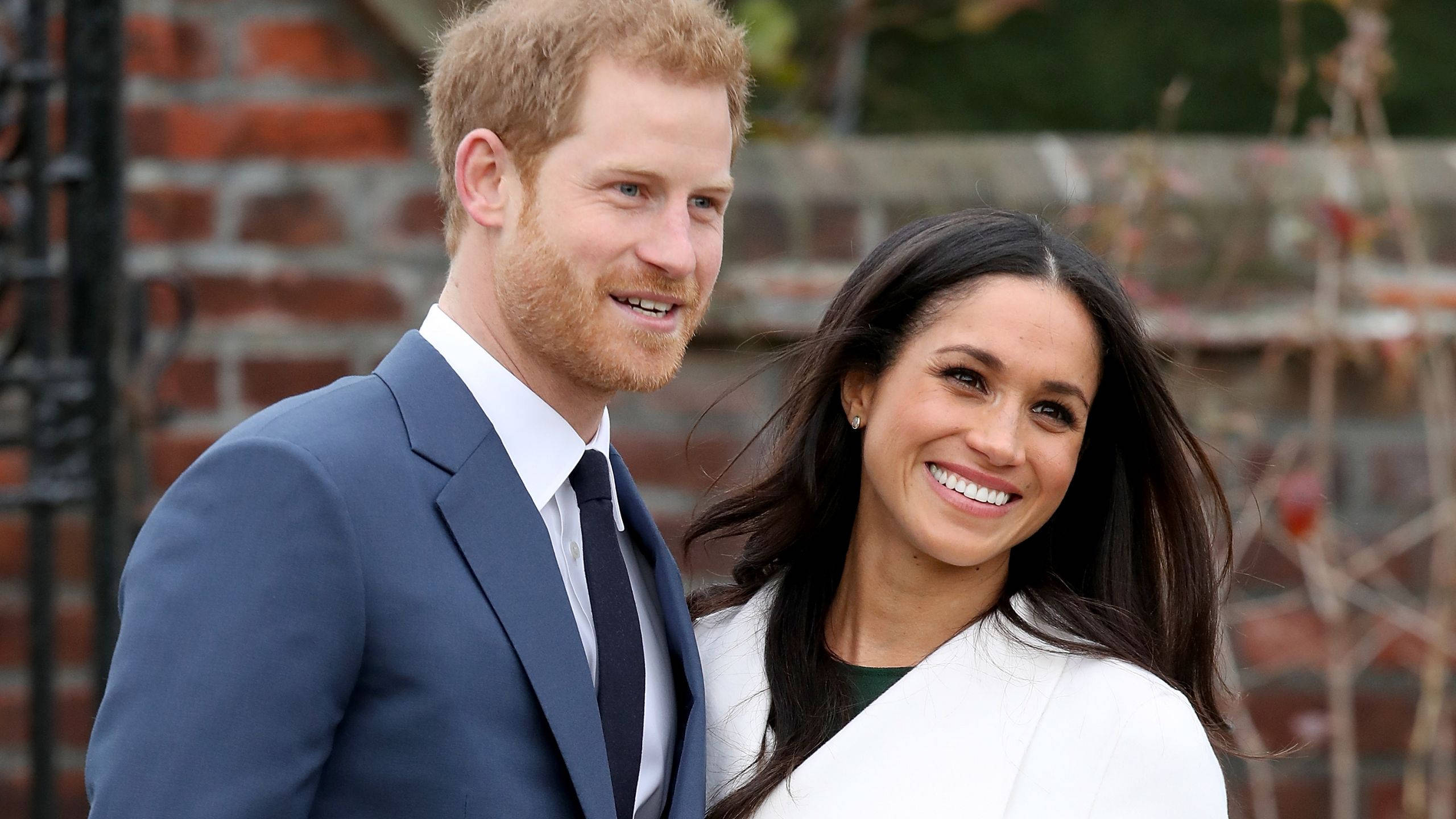 Meghan Markle 桌布 壁紙 — 哈利王子與梅根·馬克爾在紅磚牆前微笑，身後有綠植。