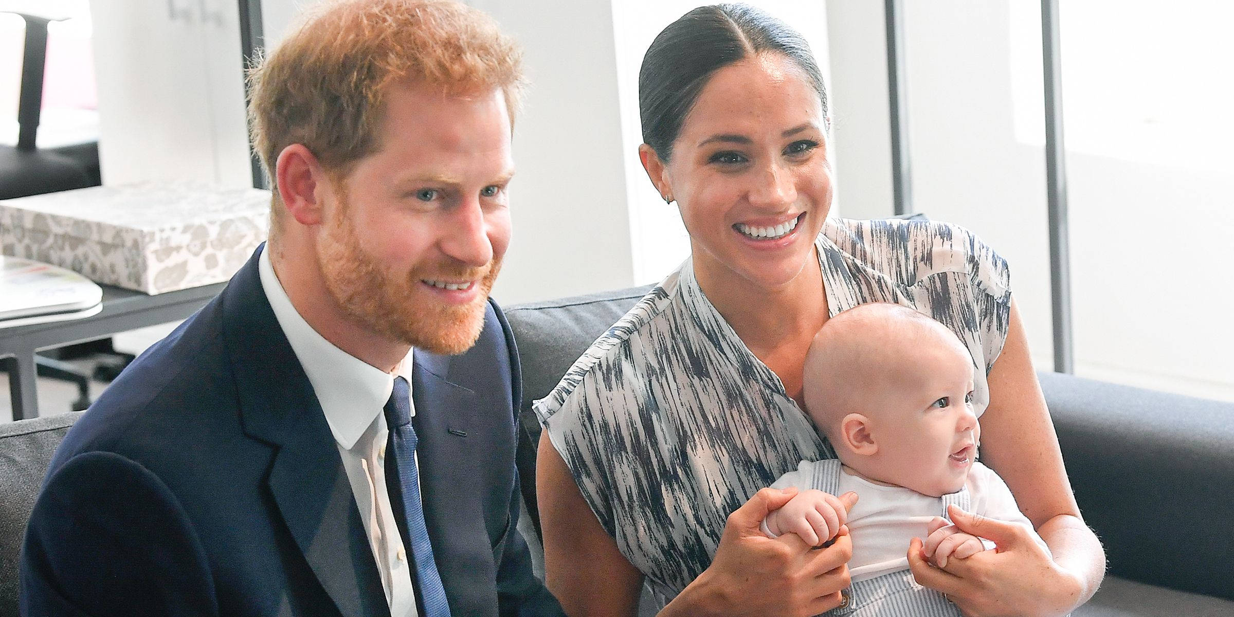 Meghan Markle 桌布 壁紙 — 哈利王子與梅根馬克爾在明亮的房間裡的現代沙發上抱著他們的兒子，溫暖地微笑著。