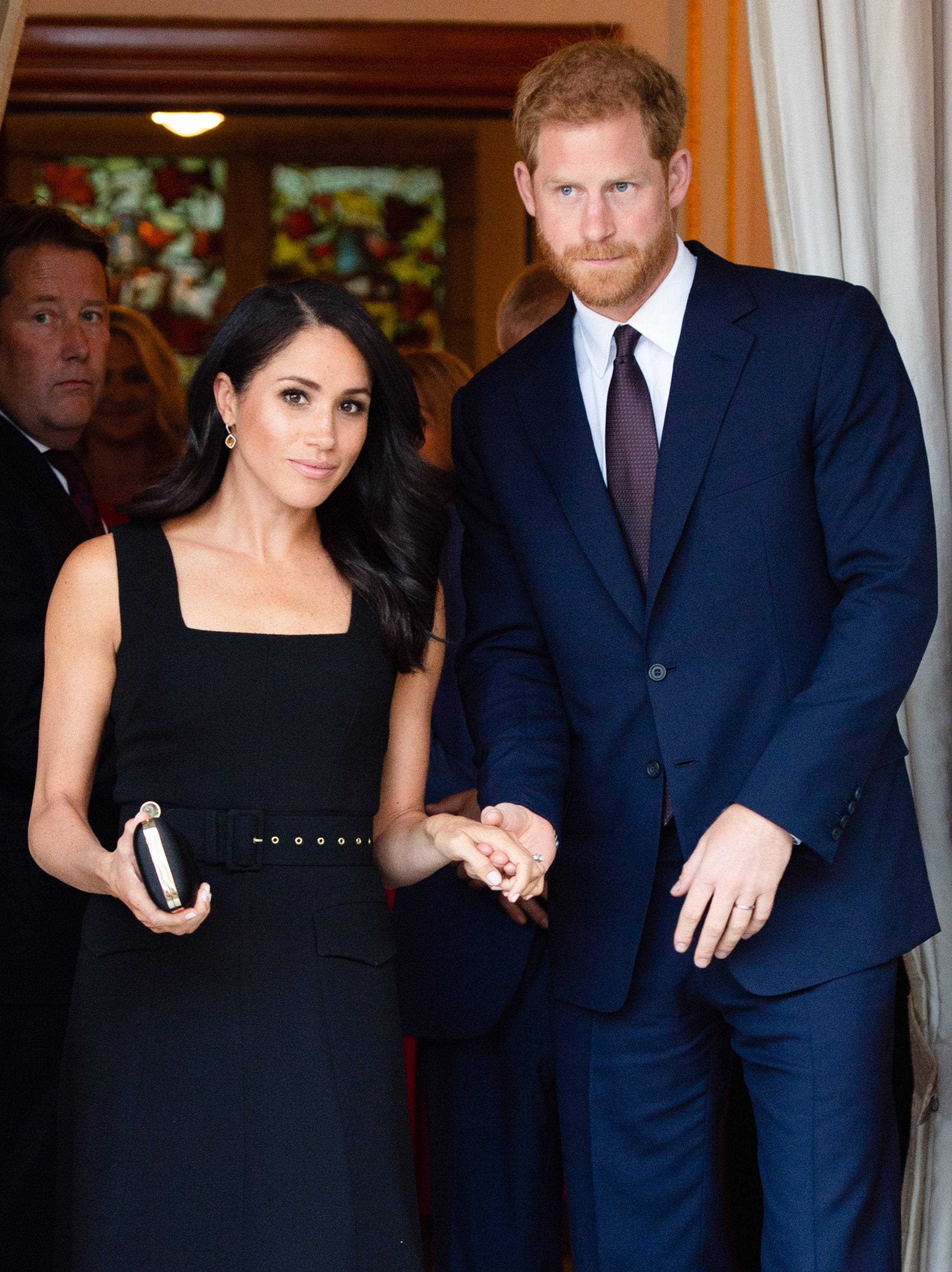 Prince 桌布 壁紙 — 哈利王子與梅根馬克爾穿著正式服裝手牽手，站在室內，身後有彩色玻璃窗。
