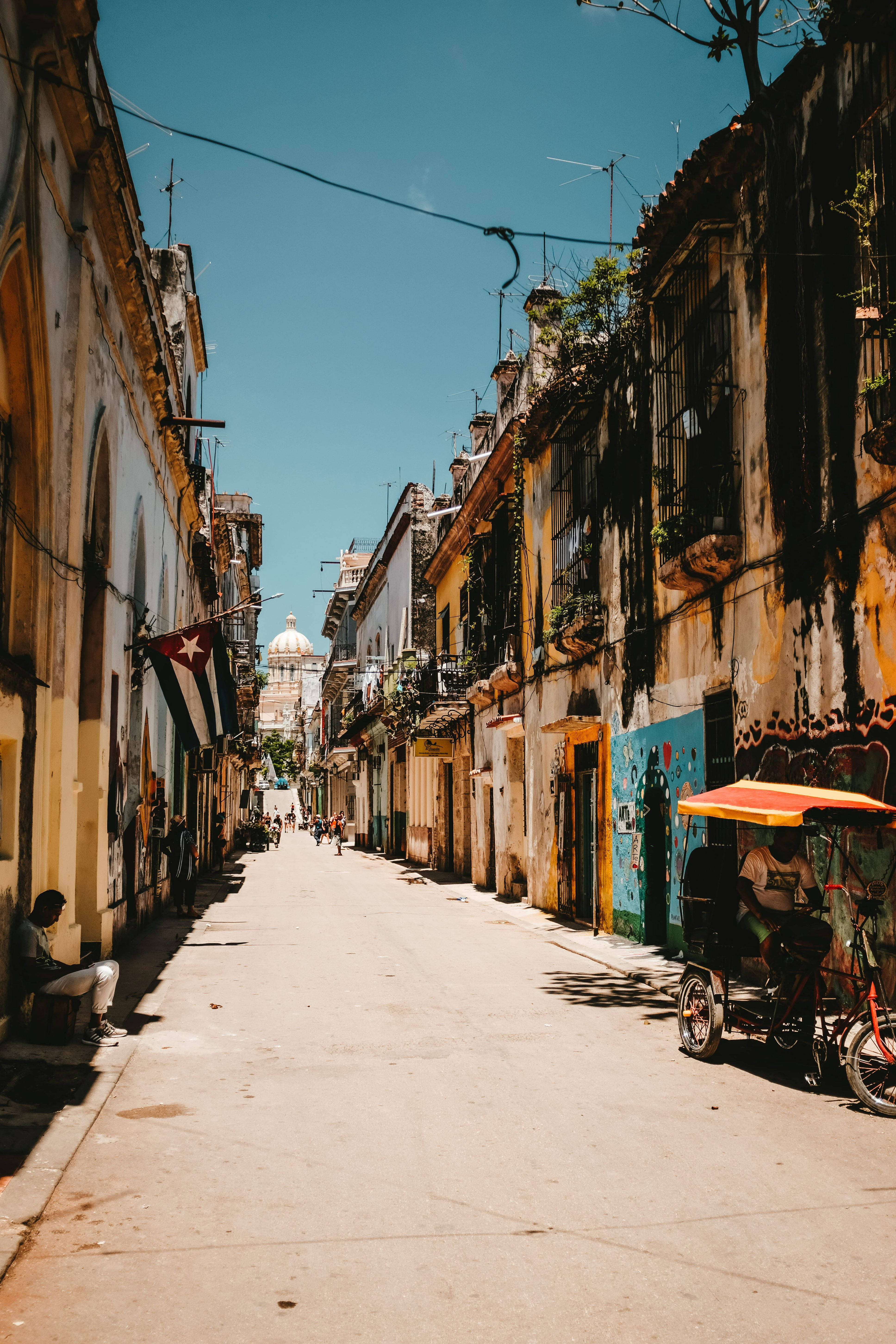 Cuban 桌布 壁紙 — 哈瓦那一條陽光普照的狹窄街道，兩旁是飽經風霜的殖民時期建築，一輛三輪車在晴朗的藍天下駛過。