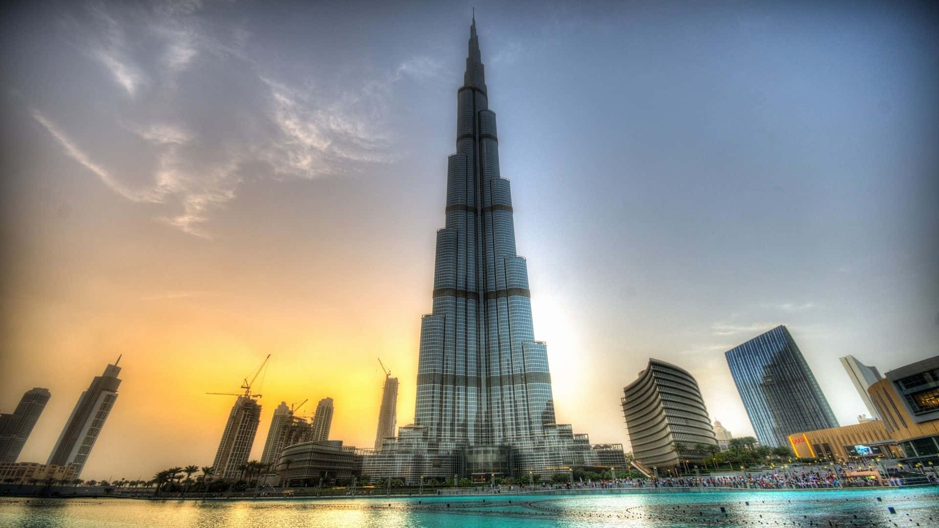 Uae 桌布 壁紙 — 哈里發塔在日落時分聳立於杜拜天際線之上，金色光芒反射在周圍的摩天大樓和水面上。