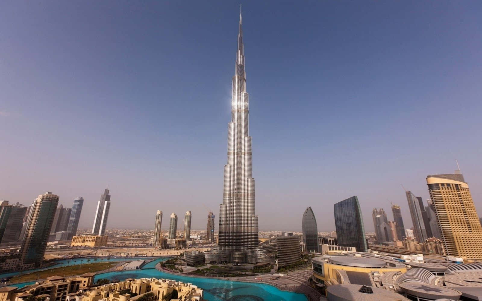 Burj Khalifa 桌布 壁紙 — 哈里發塔聳立在杜拜天際線上，其閃閃發光的尖頂刺穿清澈的藍天，下方是現代玻璃摩天大樓和藍綠色水景。