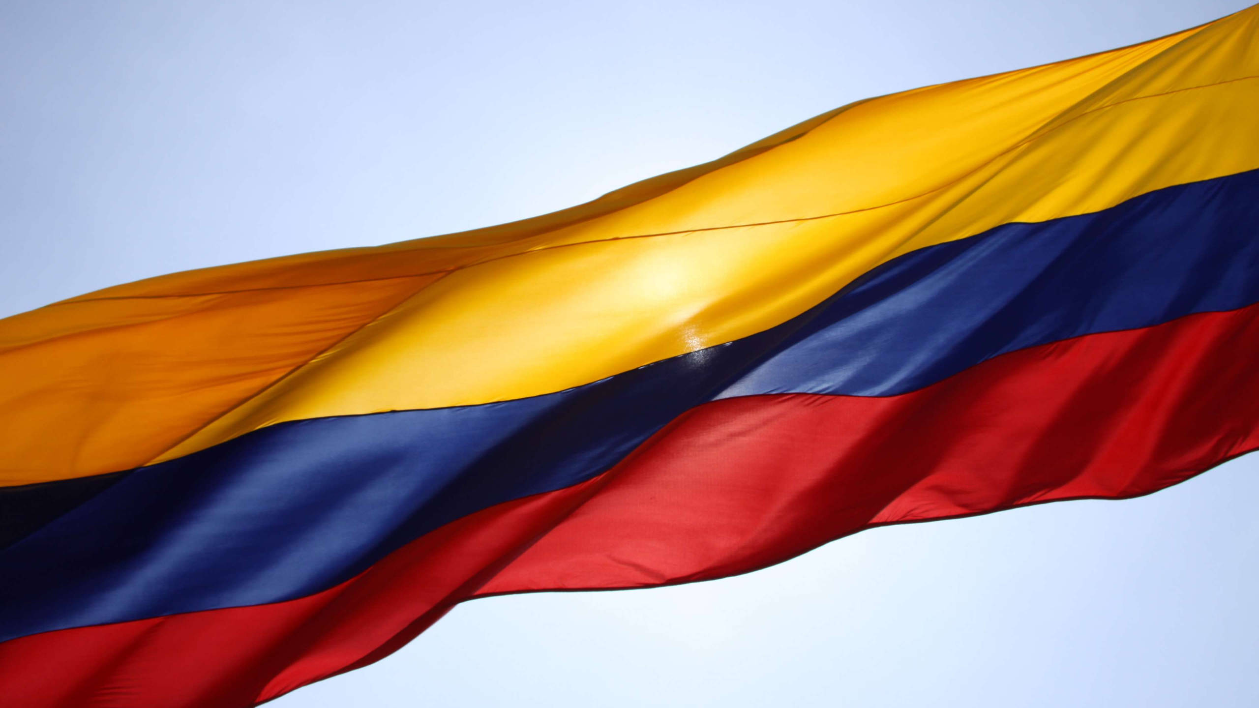 Colombia Flag 桌布 壁紙 — 哥倫比亞國旗在清澈的淡藍色天空中動態飄揚，帶有鮮豔的黃、藍、紅條紋。