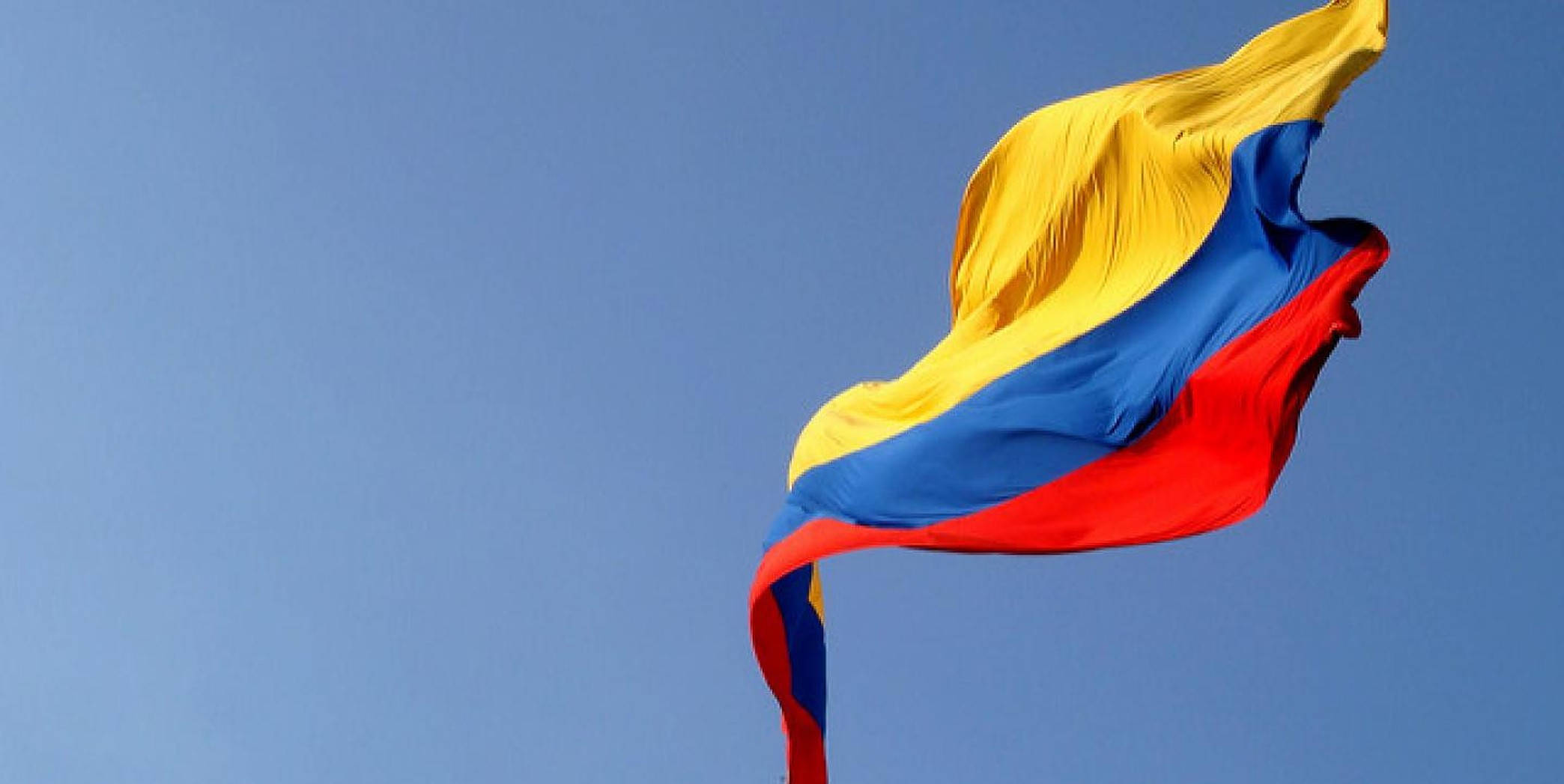 Colombia 桌布 壁紙 — 哥倫比亞國旗在清澈的藍天下動態飄揚，帶有鮮明的黃、藍、紅條紋。
