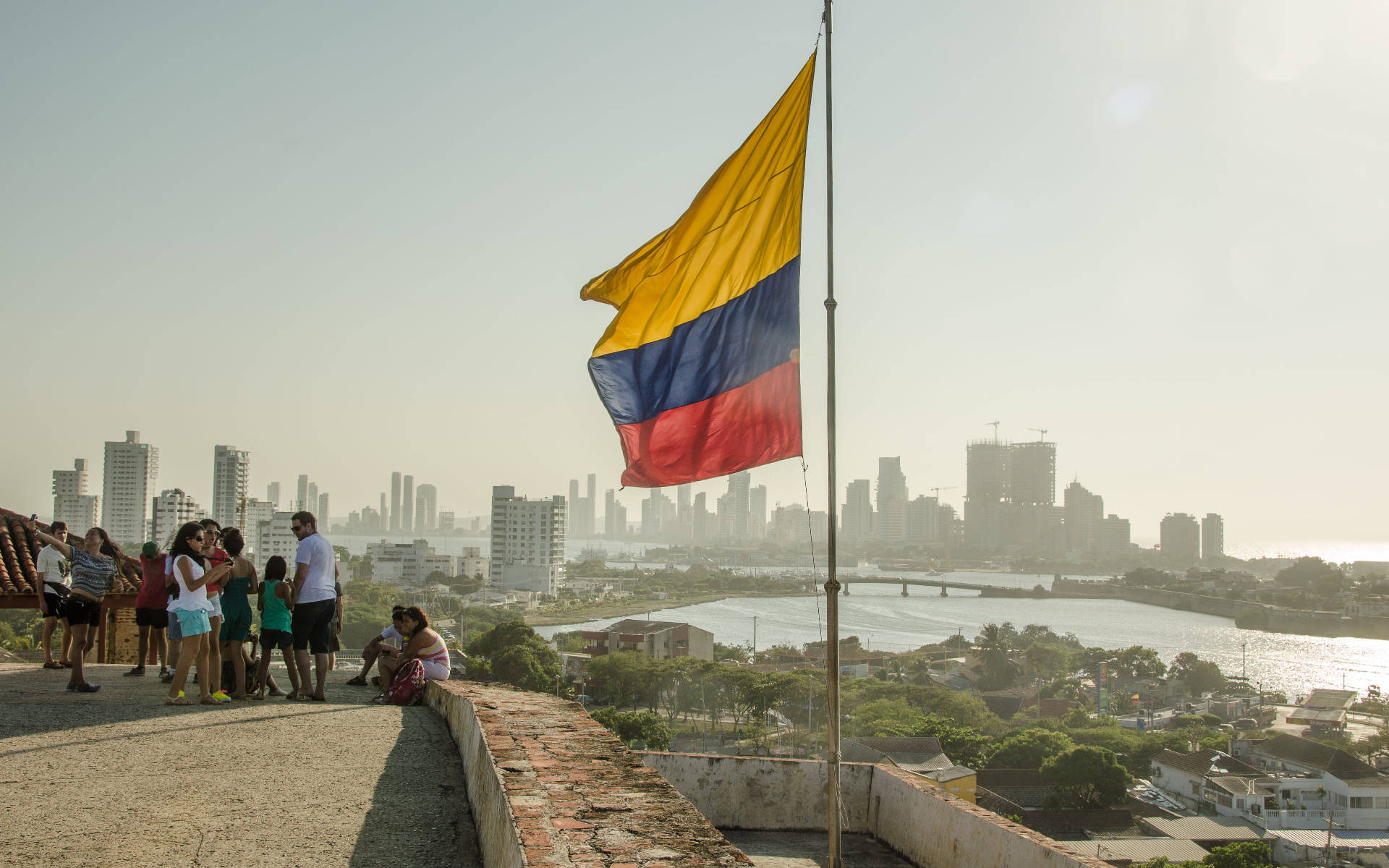 Colombia Flag 桌布 壁紙 — 哥倫比亞國旗在石頭觀景台上自豪地飄揚，遊客在黃金時刻欣賞下方朦朧的城市景觀和河流。