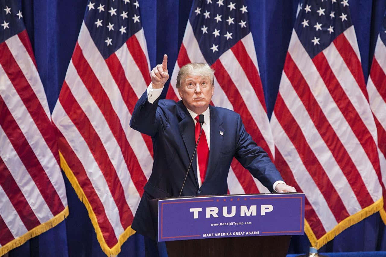 川普 桌布 壁紙 — 唐納·川普站在講台上，帶有「TRUMP」和「MAKE AMERICA GREAT AGAIN」標牌，右手食指向上指，背景為多面美國國旗。