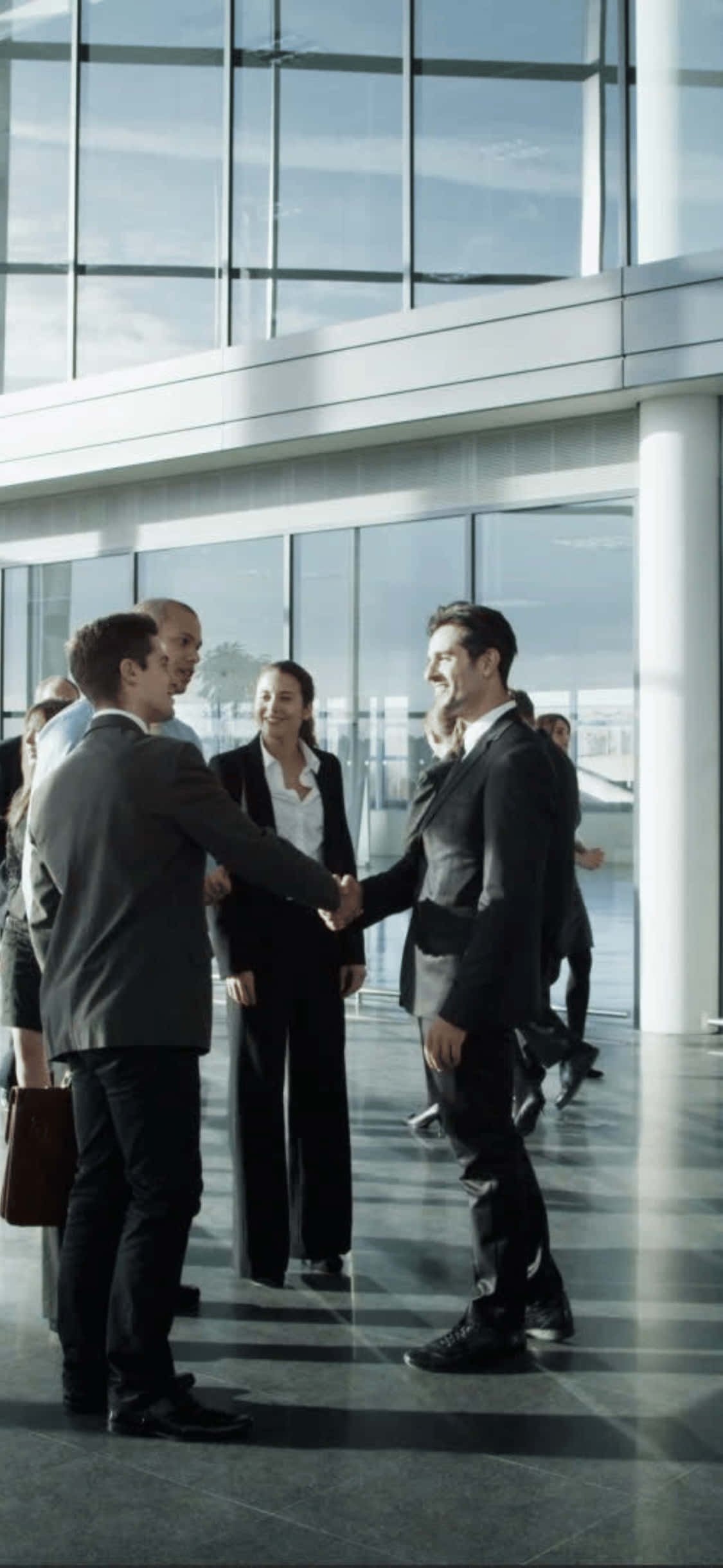 suits 桌布 壁紙 — 商務專業人士在現代玻璃牆的企業大廳中握手，自然光線透入。