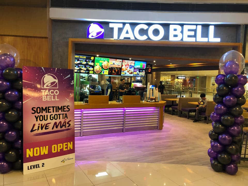 塔可鐘 桌布 壁紙 — 商場內光線明亮的Taco Bell餐廳入口，設有紫色和黑色氣球柱及「現已開業」標誌。