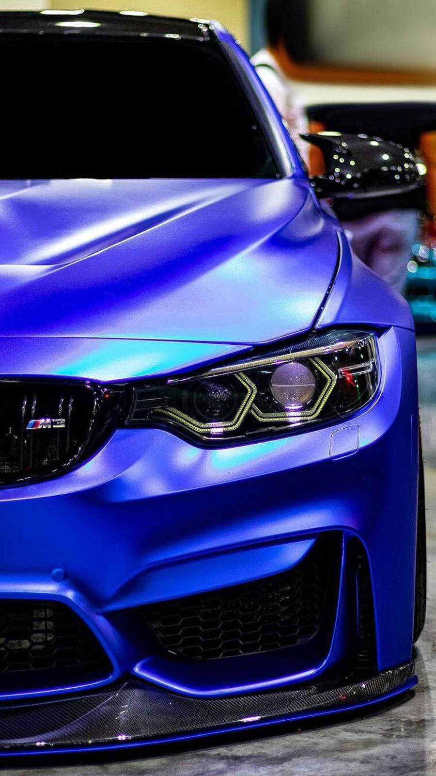 iPhone 桌布 壁紙 — 啞光藍色BMW M3的特寫，配備發光頭燈和碳纖維飾件，在明亮的室內燈光下。