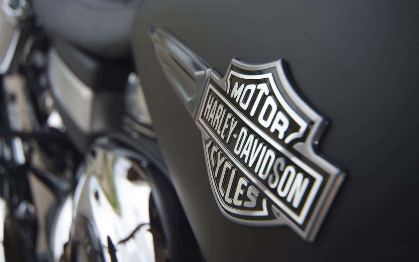 哈雷戴維森 桌布 壁紙 — 啞光黑色摩托車油箱上的鍍鉻Harley-Davidson標誌特寫，背景是模糊的鍍鉻引擎零件。