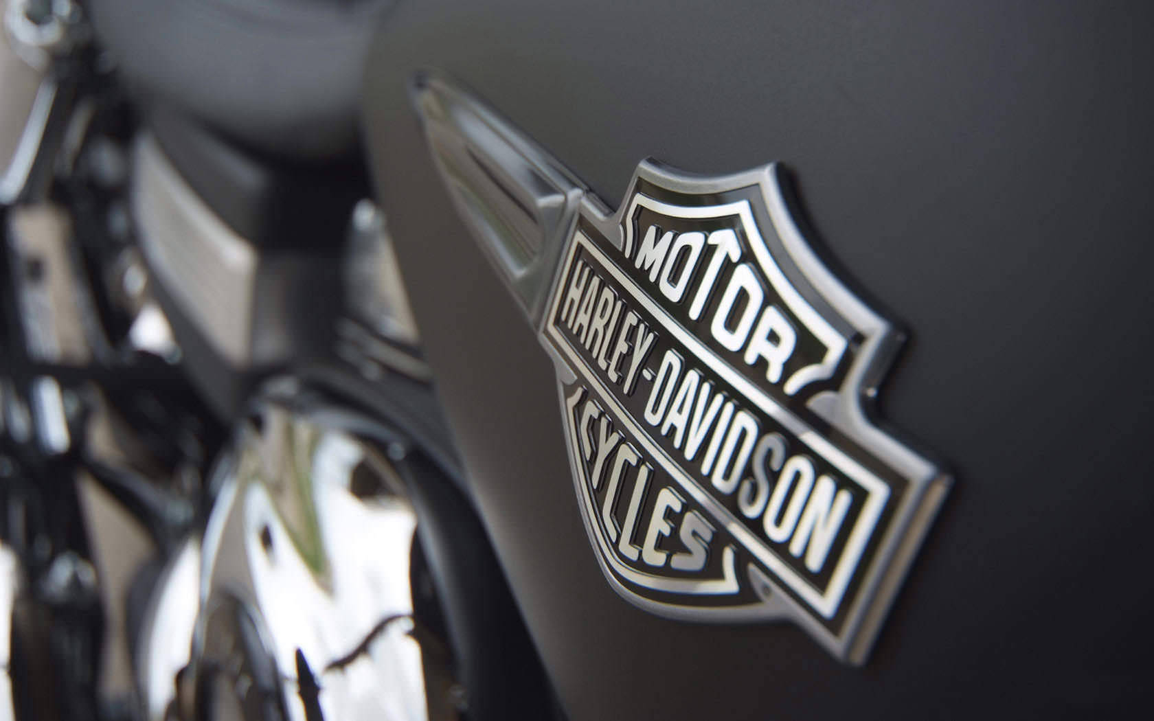 Harley 桌布 壁紙 — 啞光黑色機車油箱上的鍍鉻 Harley-Davidson 標誌特寫，背景有模糊的鍍鉻引擎零件。