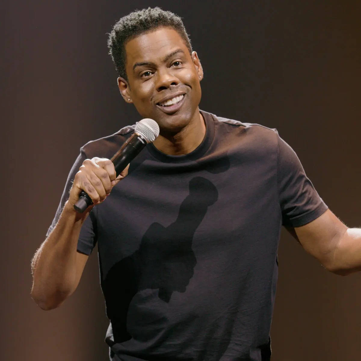 Chris Rock 桌布 壁紙 — 喜劇演員克里斯·洛克在溫暖的聚光燈下在舞台上拿著麥克風微笑。