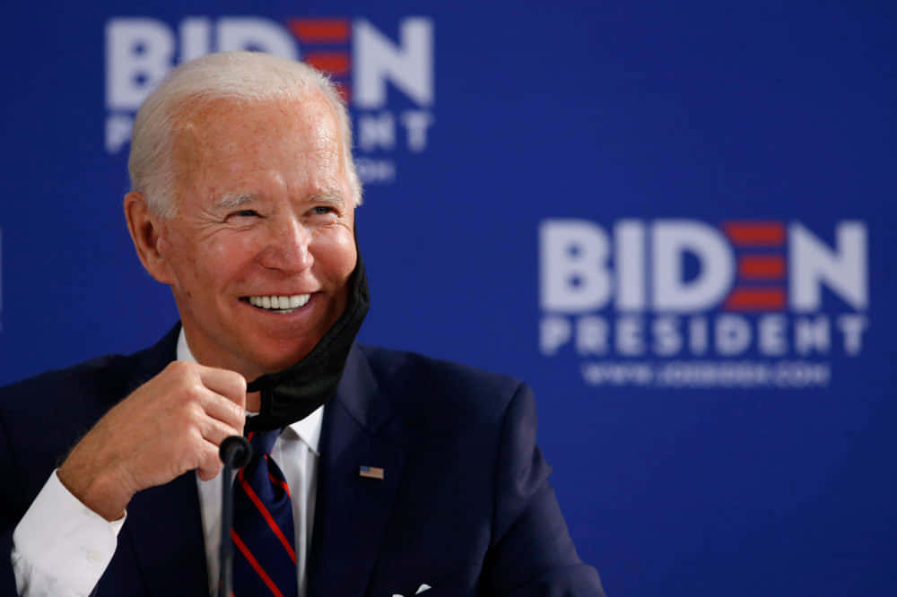 喬·拜登 桌布 壁紙 — 喬·拜登在藍色「Biden President」背景的競選活動中，手持口罩，露出燦爛笑容。