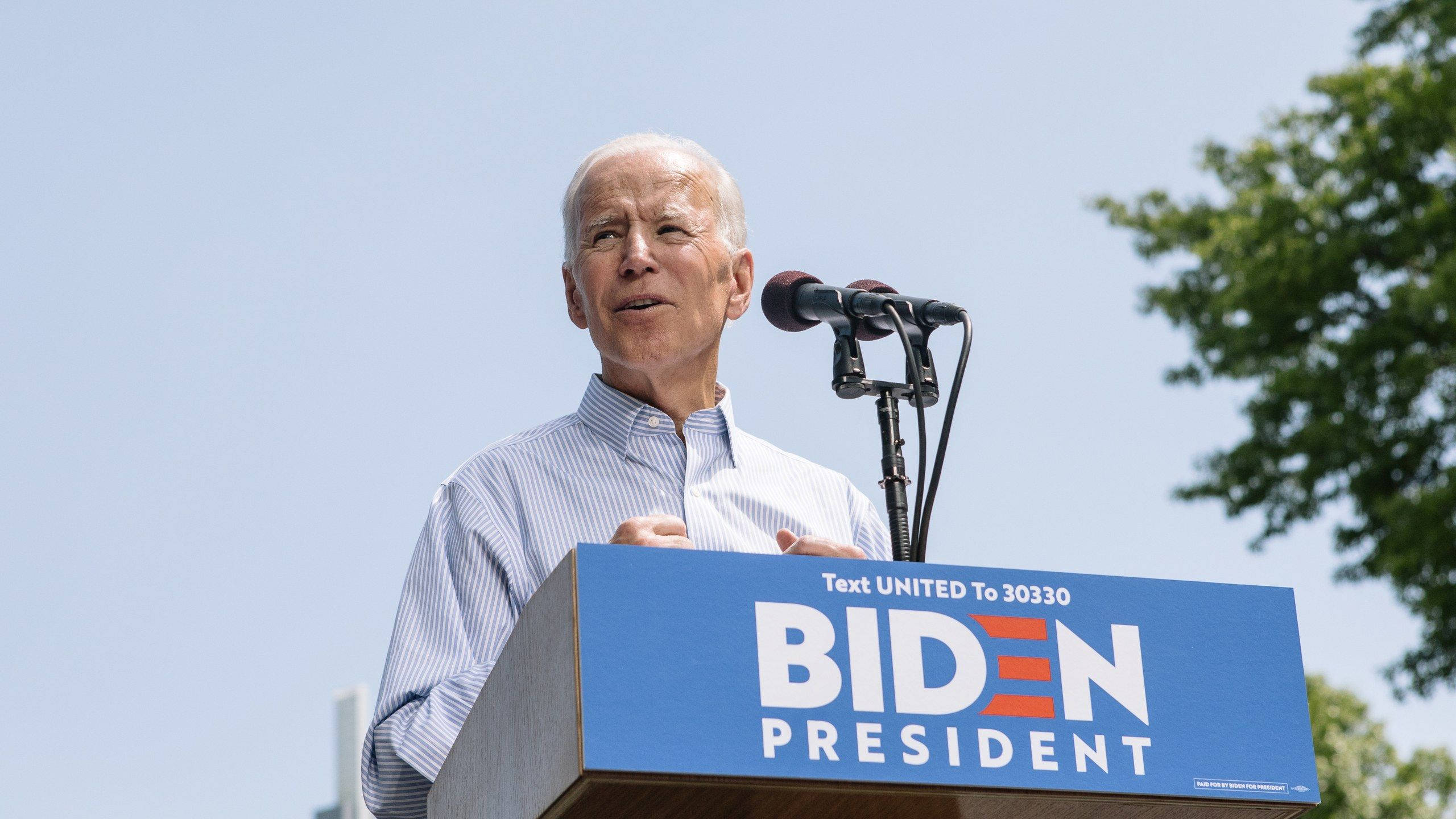 Biden 桌布 壁紙 — 喬·拜登在藍色講台後方，講台上有「Biden President」字樣，在晴朗天空下進行競選集會演講。