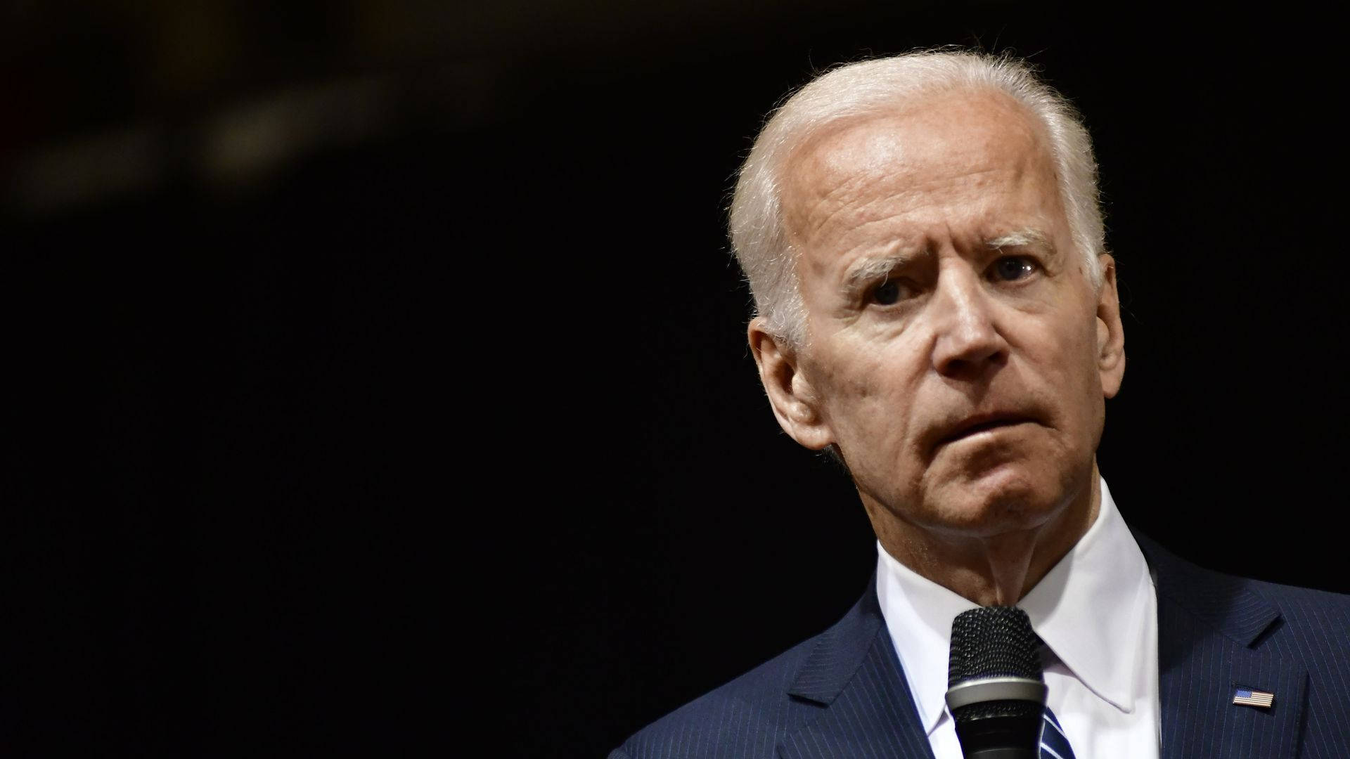 Biden 桌布 壁紙 — 喬·拜登身穿深色西裝，在黑色背景前對著麥克風講話的特寫肖像。