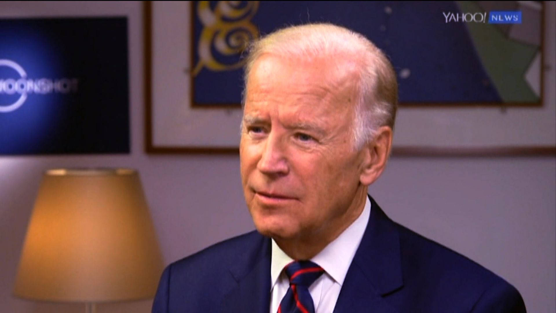 Biden 桌布 壁紙 — 喬·拜登身穿深色西裝和條紋領帶，坐在光線柔和的室內環境中，身後有裱框藝術品和一盞燈的特寫。