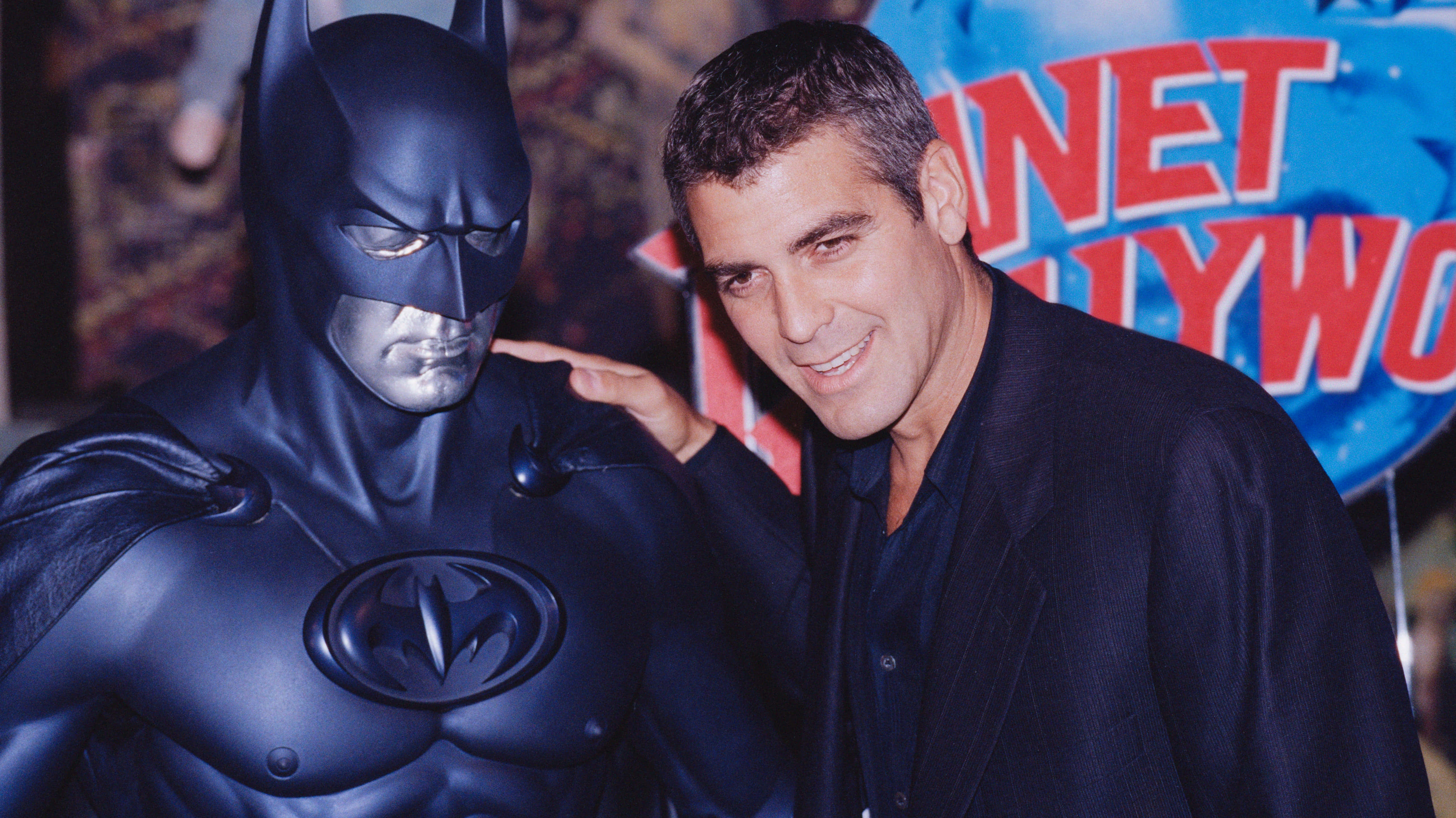 Bat-suit 桌布 壁紙 — 喬治克隆尼在 Planet Hollywood 活動中，在閃亮的蝙蝠俠雕像旁溫暖地微笑。