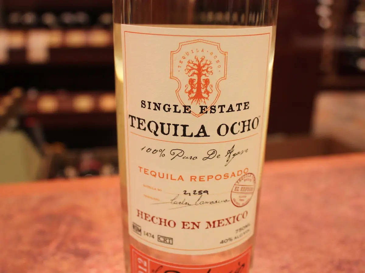 bottle 桌布 壁紙 — 單一莊園龍舌蘭酒瓶的特寫，標籤為「Tequila Ocho」，放在木吧檯上，背景有模糊的架子。