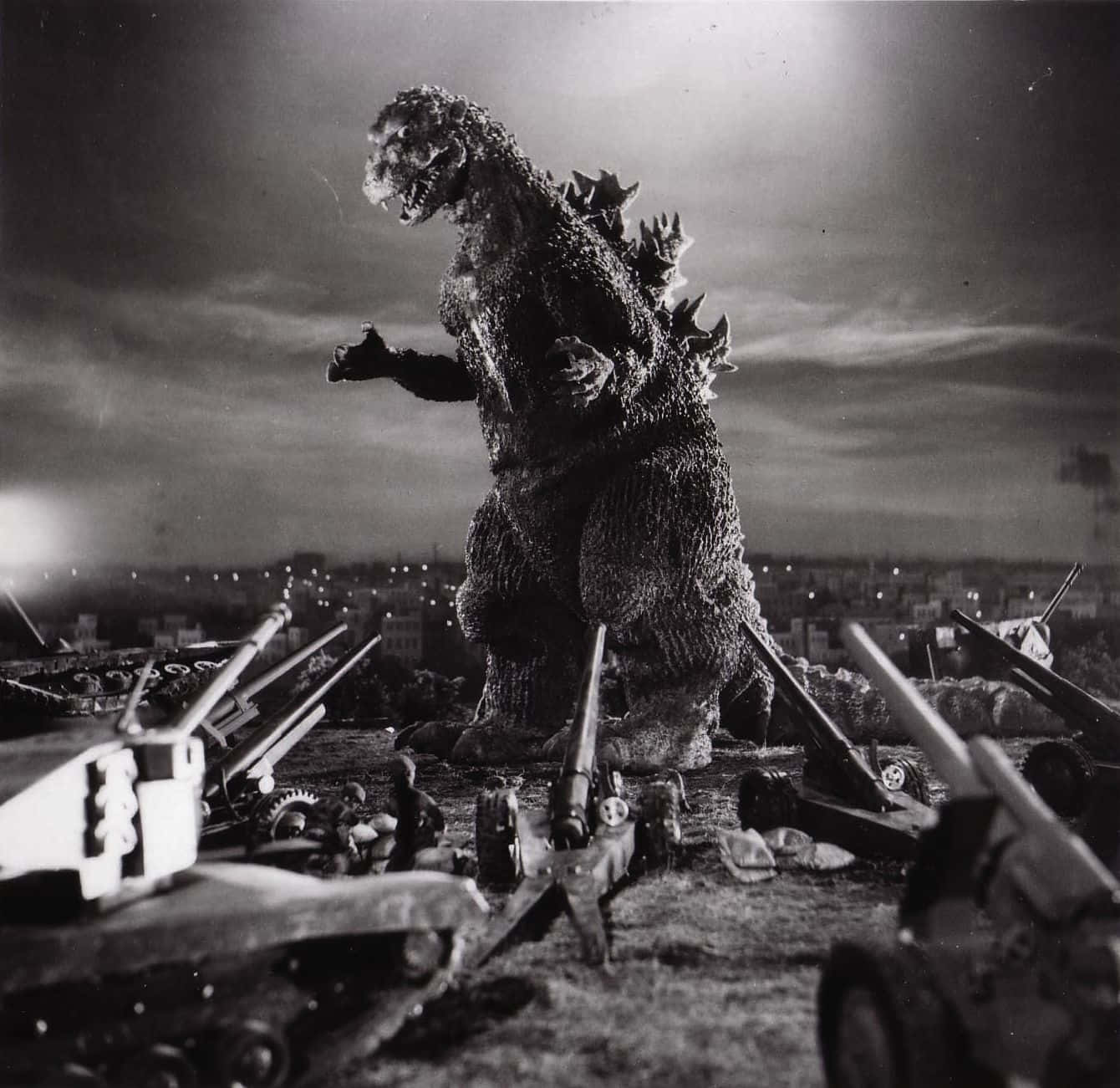 Godzilla 1954 桌布 壁紙 — 單色哥斯拉站在一個被摧毀坦克的戰場上，夜空多雲，遠處有城市燈光。