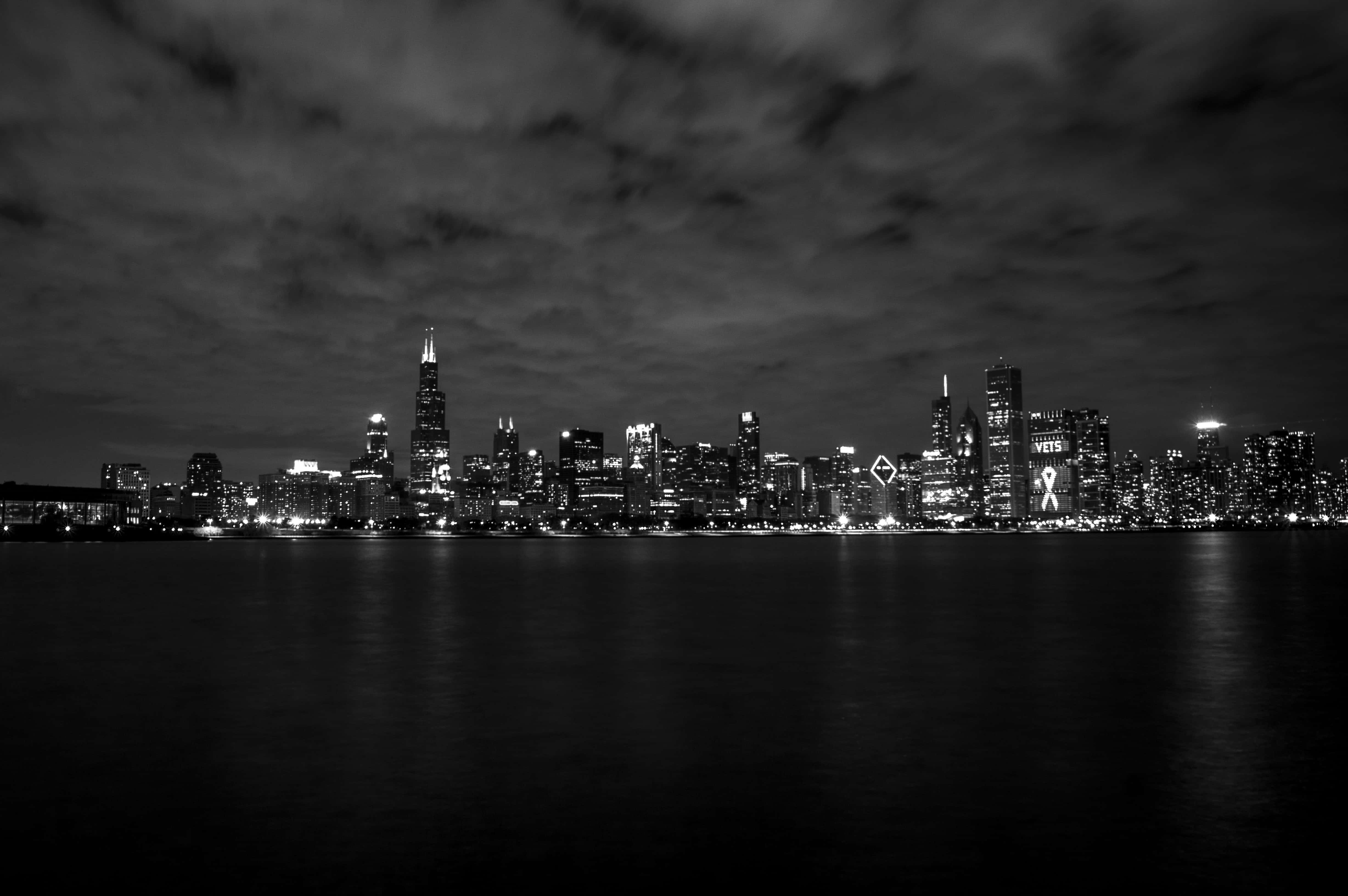 黑白 桌布 壁紙 — 單色城市天際線夜景，發光的摩天大樓倒映在平靜的水面上，天空多雲。