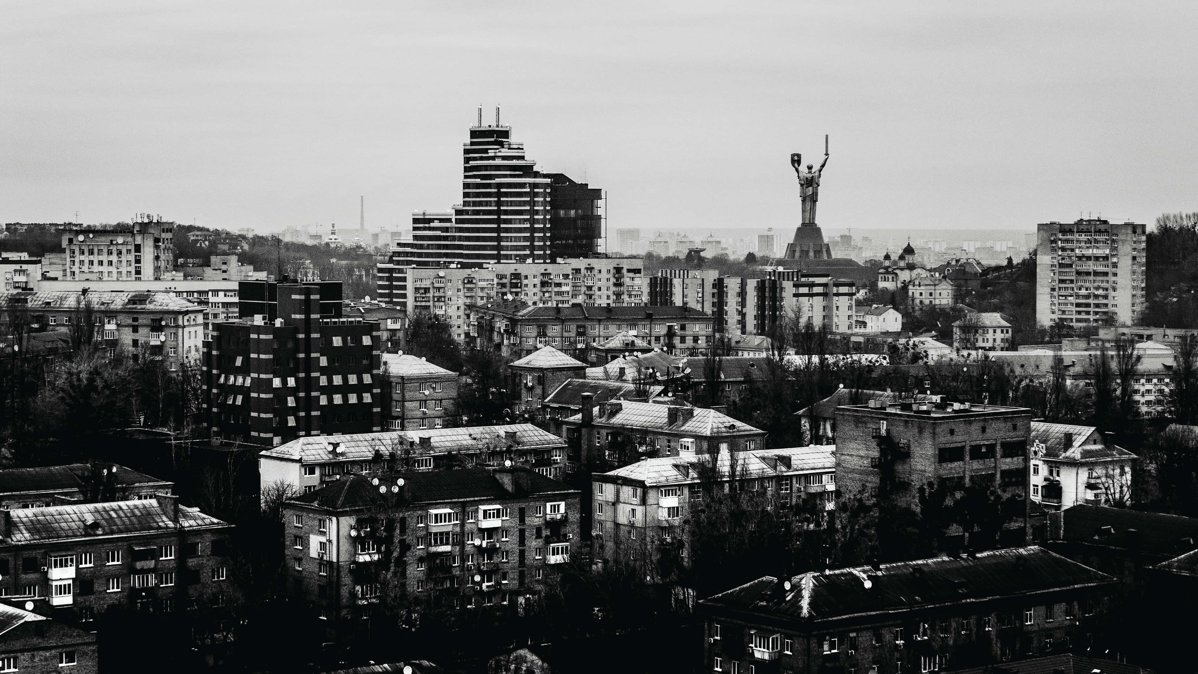 Black And White City 桌布 壁紙 — 單色城市景觀，一座高聳的舉臂人物雕像矗立在密集的住宅和現代建築之上。