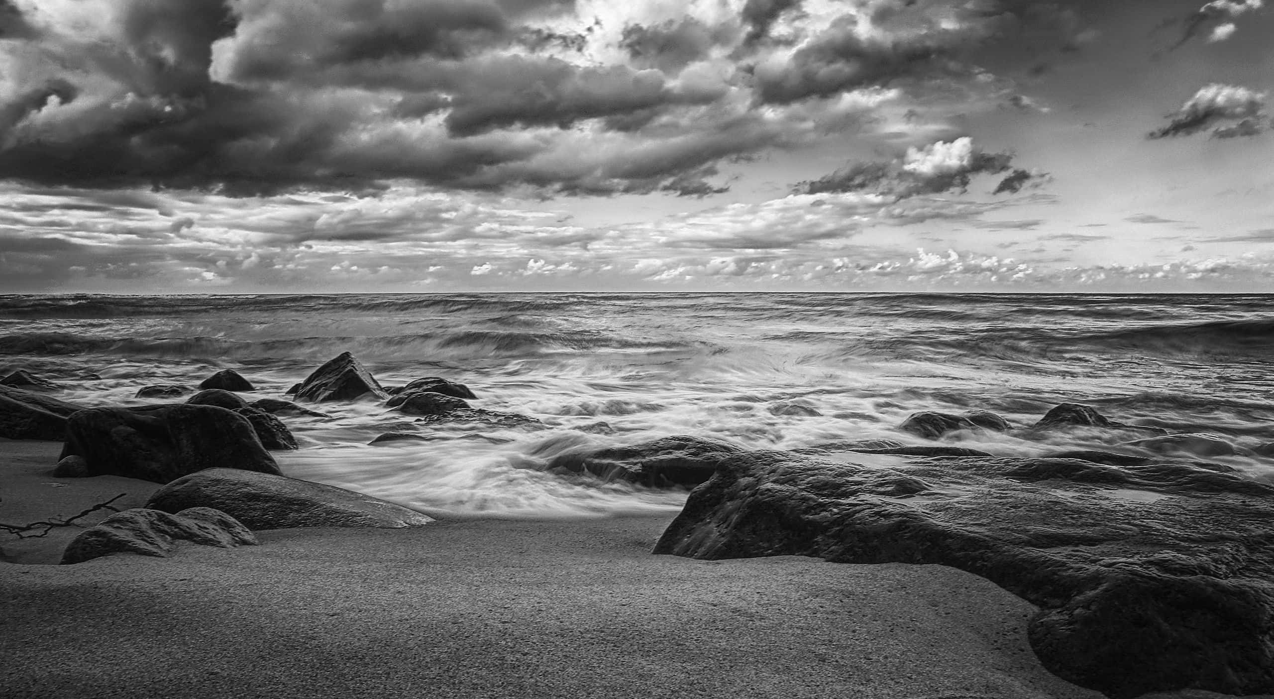 Black And White Ocean 桌布 壁紙 — 單色波浪在戲劇性的多雲天空下衝擊著海岸線的堅固岩石。