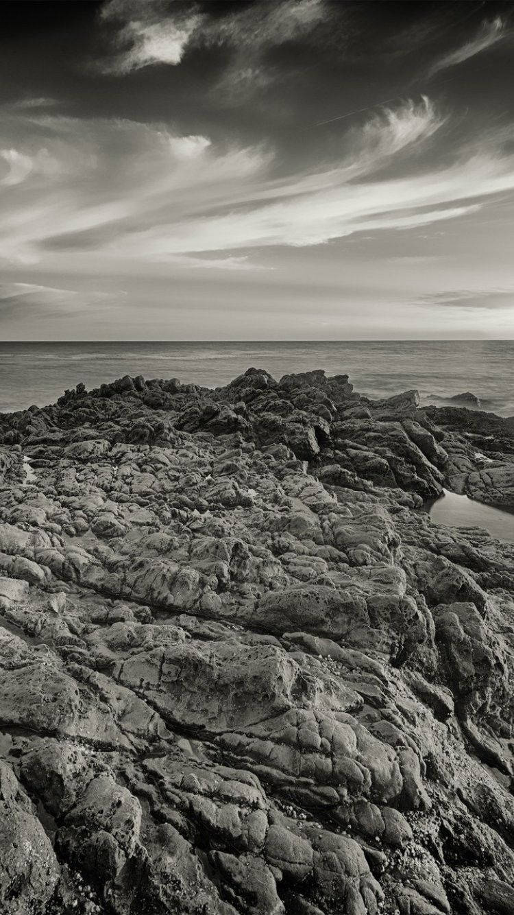 malibu iPhone 桌布 壁紙 — 單色海岸場景，紋理岩石前景，平靜海面上方動態雲層天空。