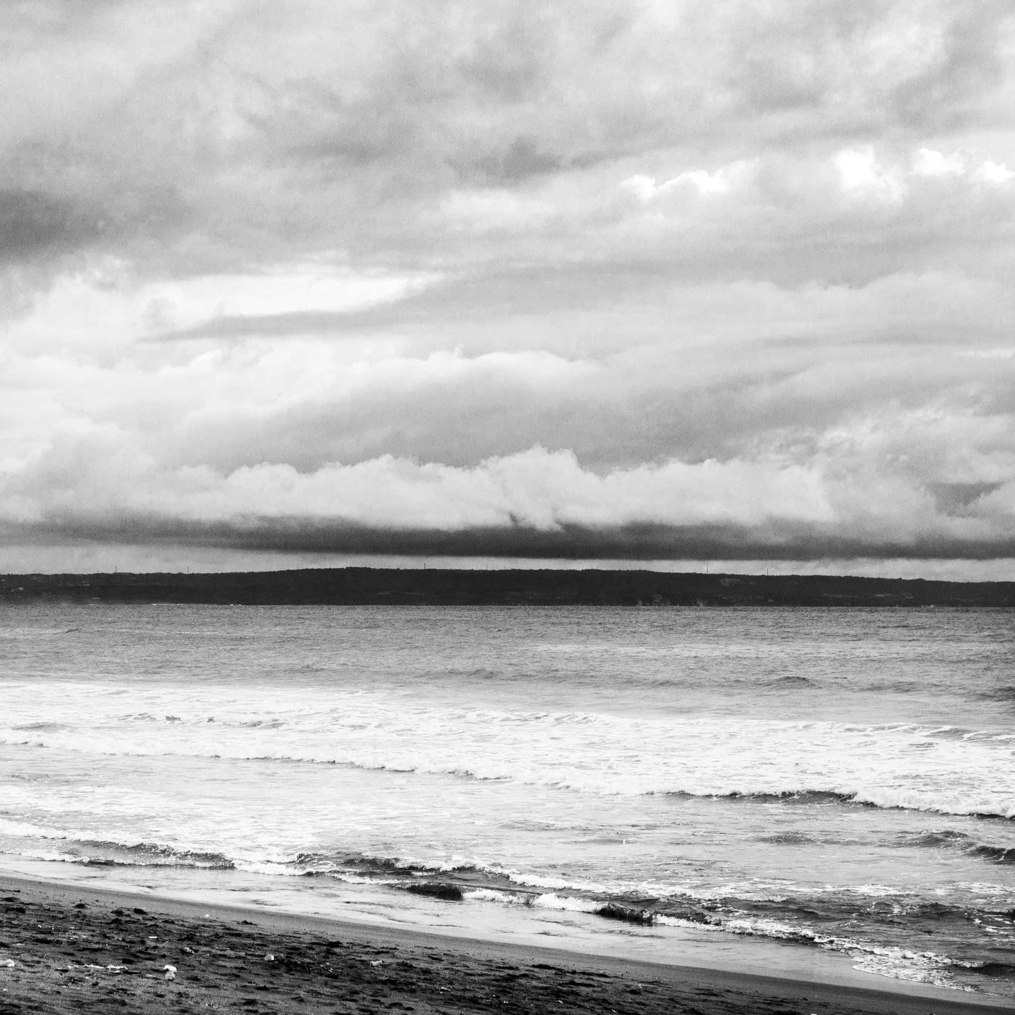 Black And White Beach 桌布 壁紙 — 單色海岸場景，遠處海岸線上有翻騰的海浪和戲劇性的雲層。