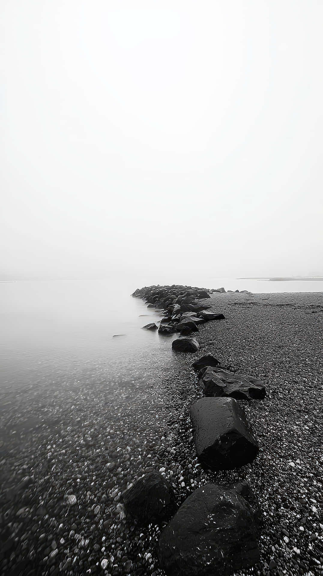 Minimalist black and white seascape with rocks fading into mist 壁紙 — 單色海岸線，深色岩石和鵝卵石在陰天天空下延伸至霧氣瀰漫的水面。