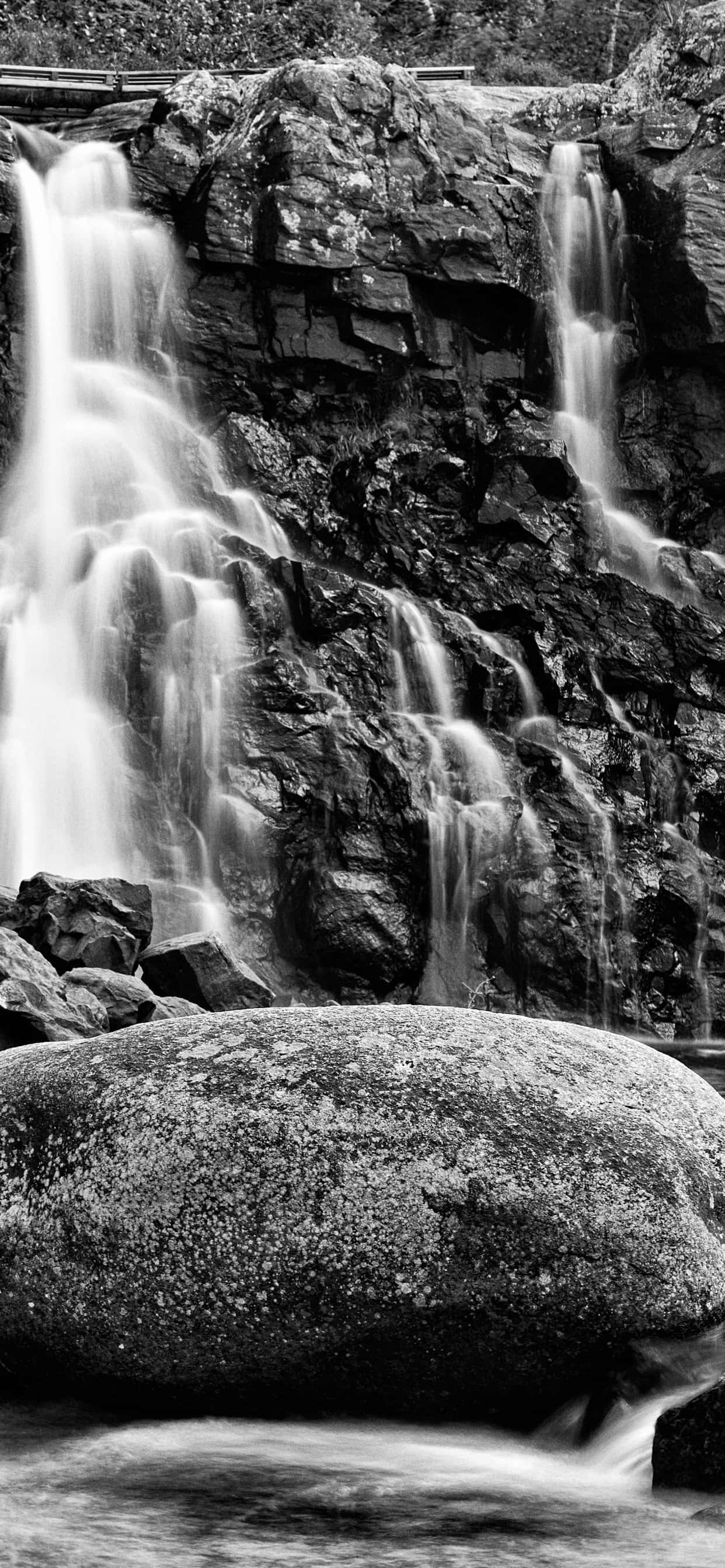 Black And White iPhone 桌布 壁紙 — 單色瀑布從崎嶇的懸崖傾瀉而下，前景有一塊大石頭。