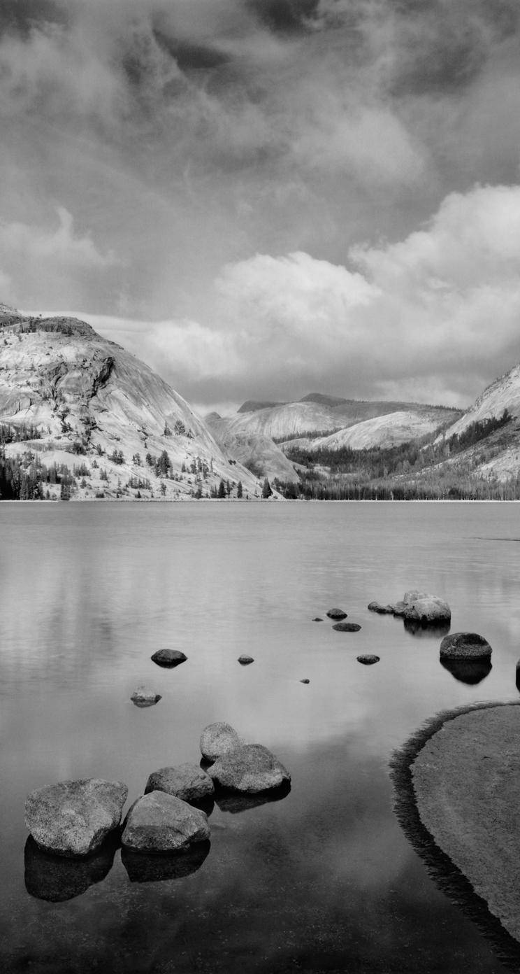 Black And White iPhone 桌布 壁紙 — 單色調的山湖，平靜的水面映照著多雲的天空，前景散落著巨石。