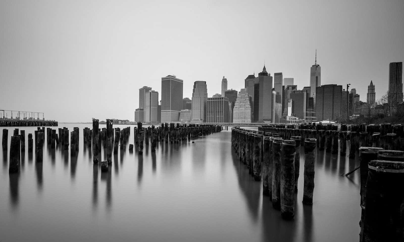 Black And White Architecture 桌布 壁紙 — 單色長時間曝光拍攝紐約市天際線倒映在平靜的水面上，並以風化的木樁框住。