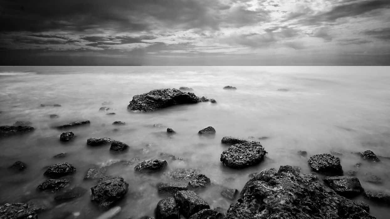 Black And White Nature 桌布 壁紙 — 單色長曝光海景，平滑的水流繞過紋理岩石，在戲劇性的多雲天空下。