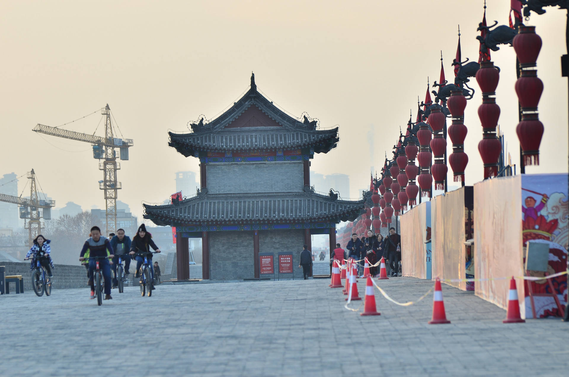 Xian 桌布 壁紙 — 單車騎士騎過一座掛滿紅燈籠的傳統中式牌樓，背景是帶有建築起重機的朦朧城市天際線。